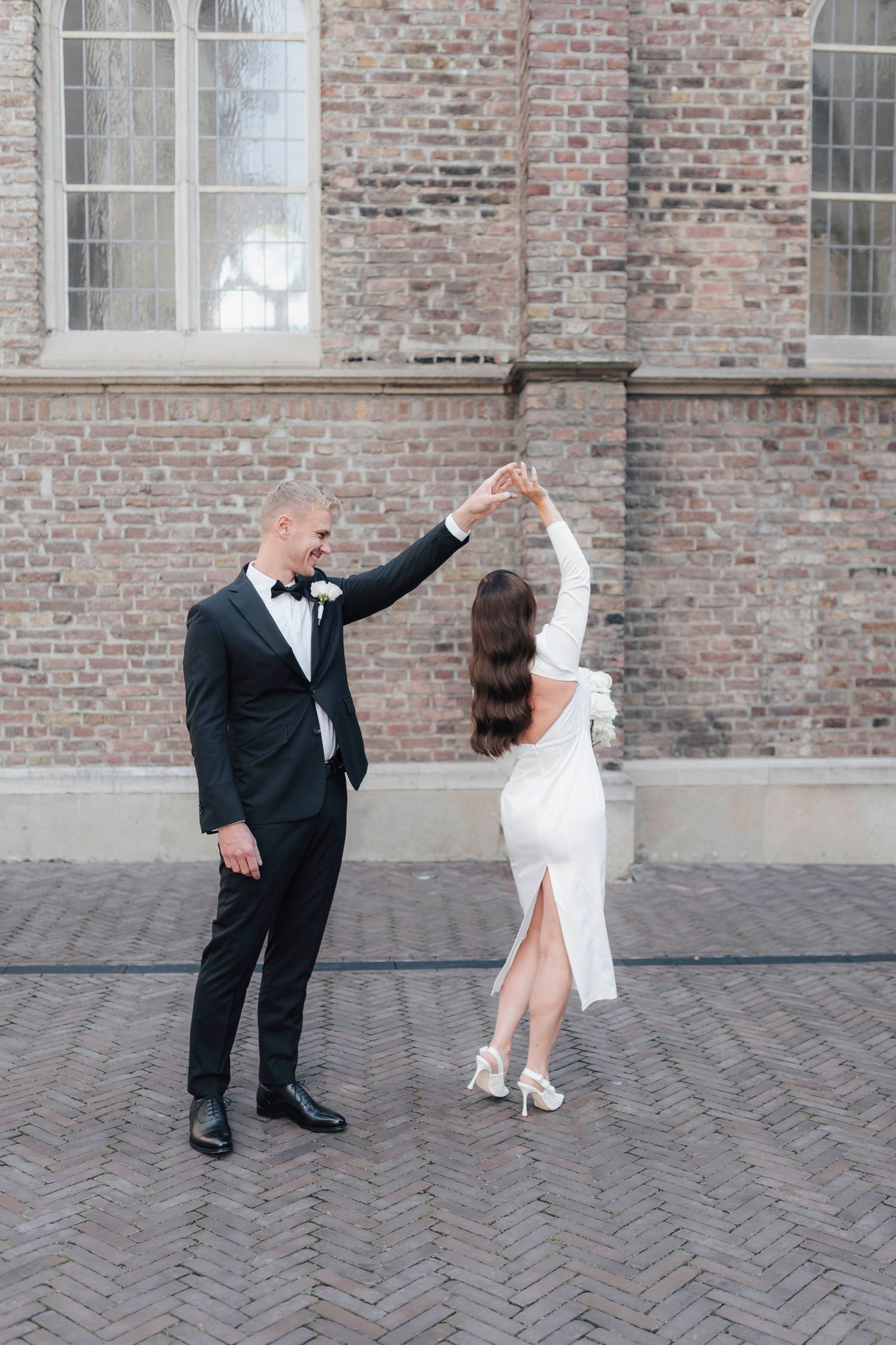 Groom in black tuxedo twirling bride in white dress outside historic brick building on cobblestone.