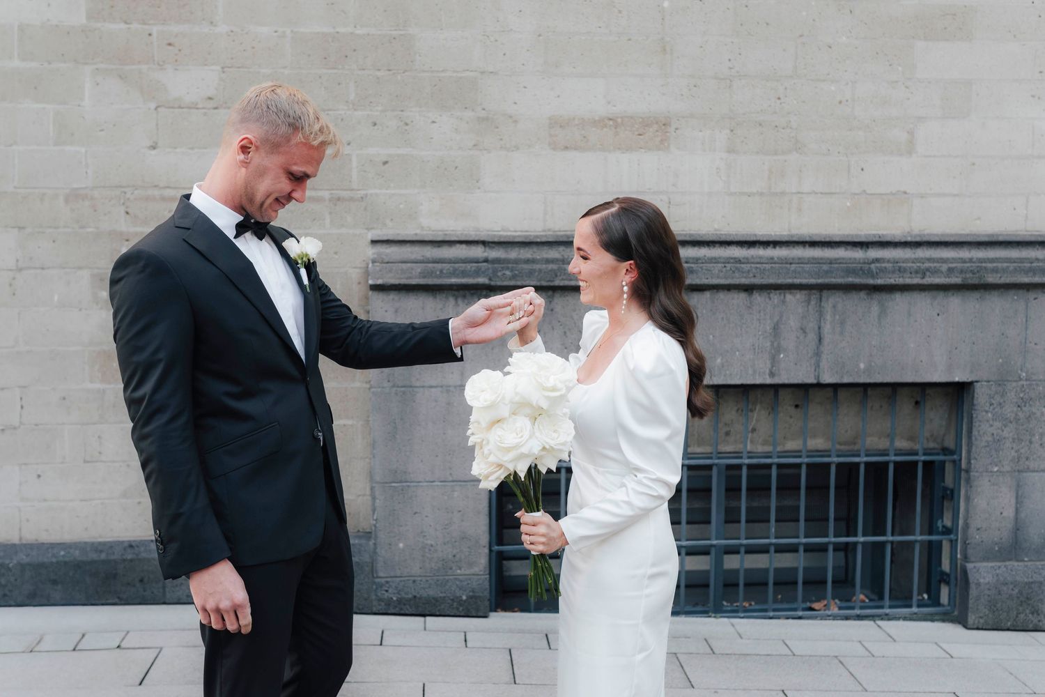 Bride and groom sharing a tender moment outdoors, she in a white gown holding florals, he in a black tuxedo.
