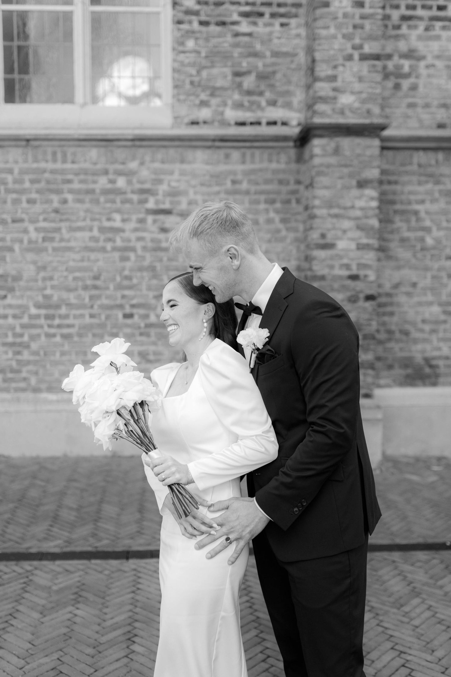 Black and white wedding photo of bride and groom embracing before a brick wall, bride holding white bouquet.