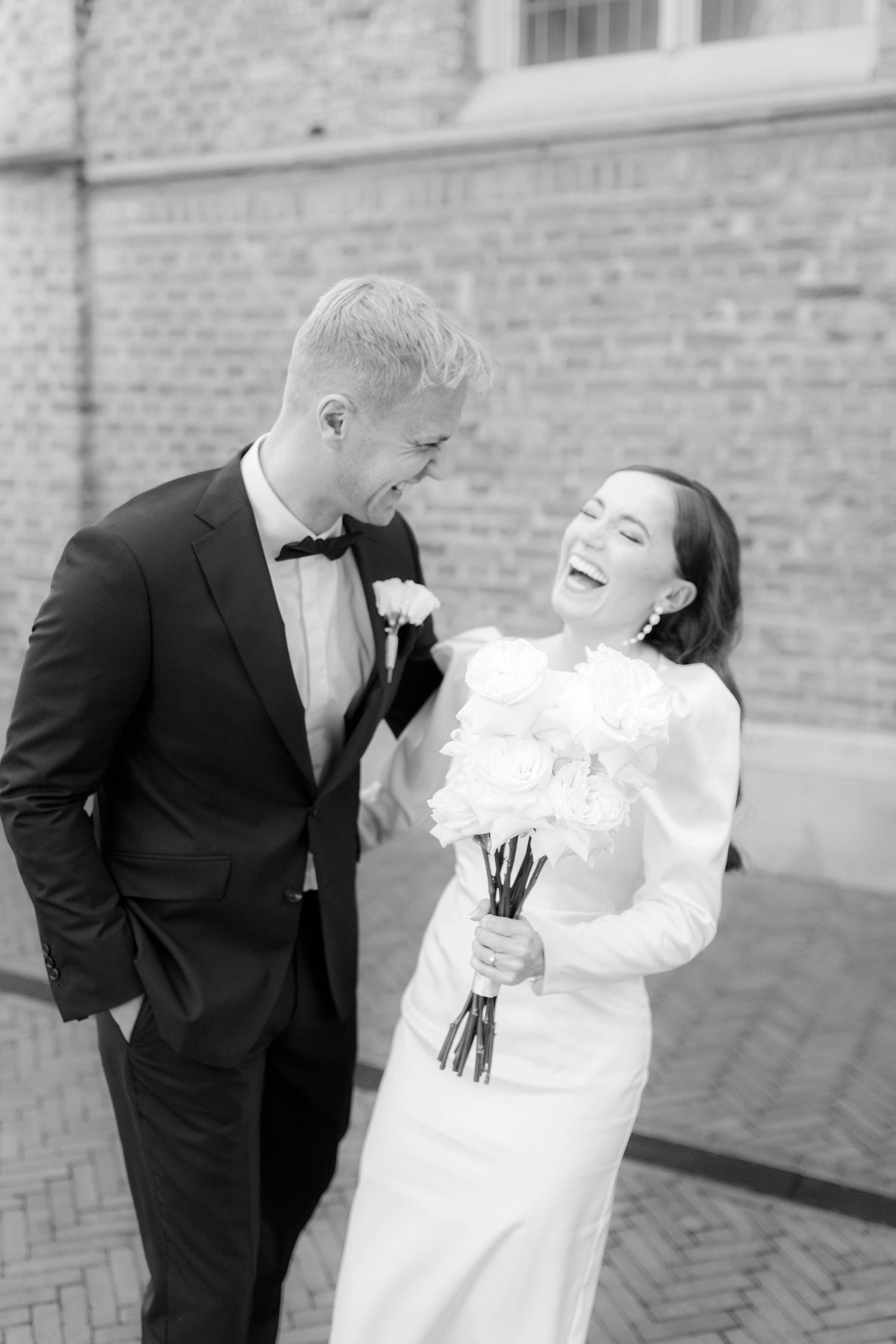 Black and white photo of a laughing bride holding white roses beside a smiling groom in a tuxedo outdoors.