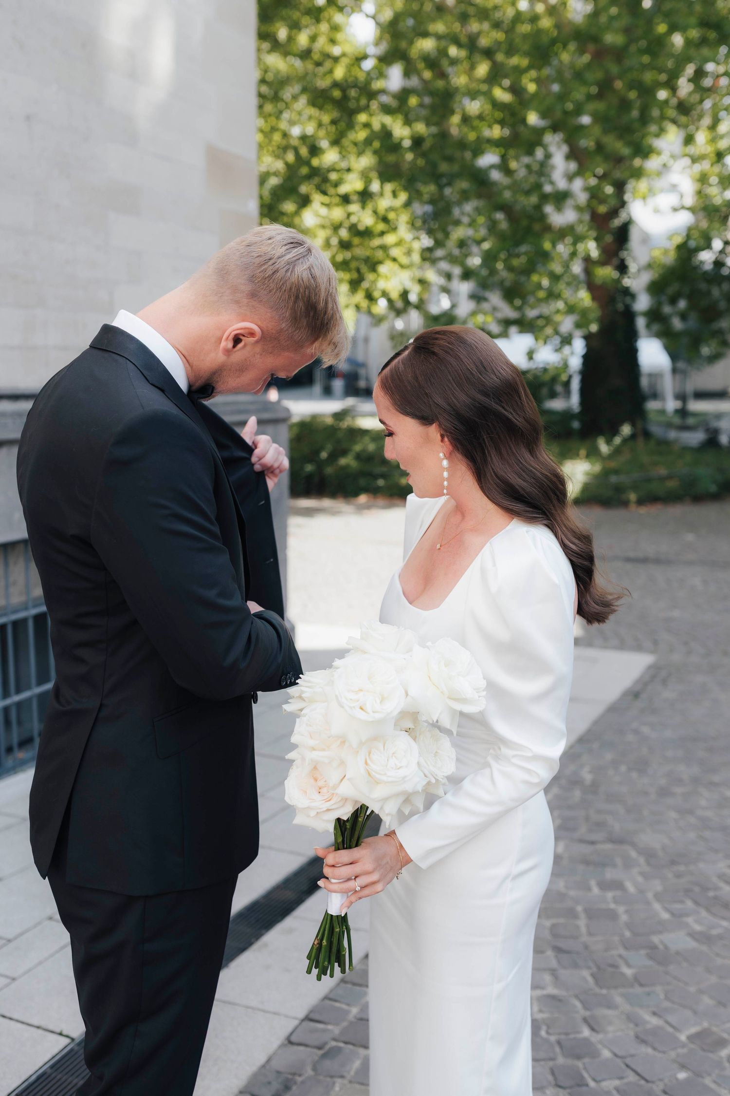 Bride in white gown holding white roses bouquet stands with groom in black tuxedo outdoors on wedding day.