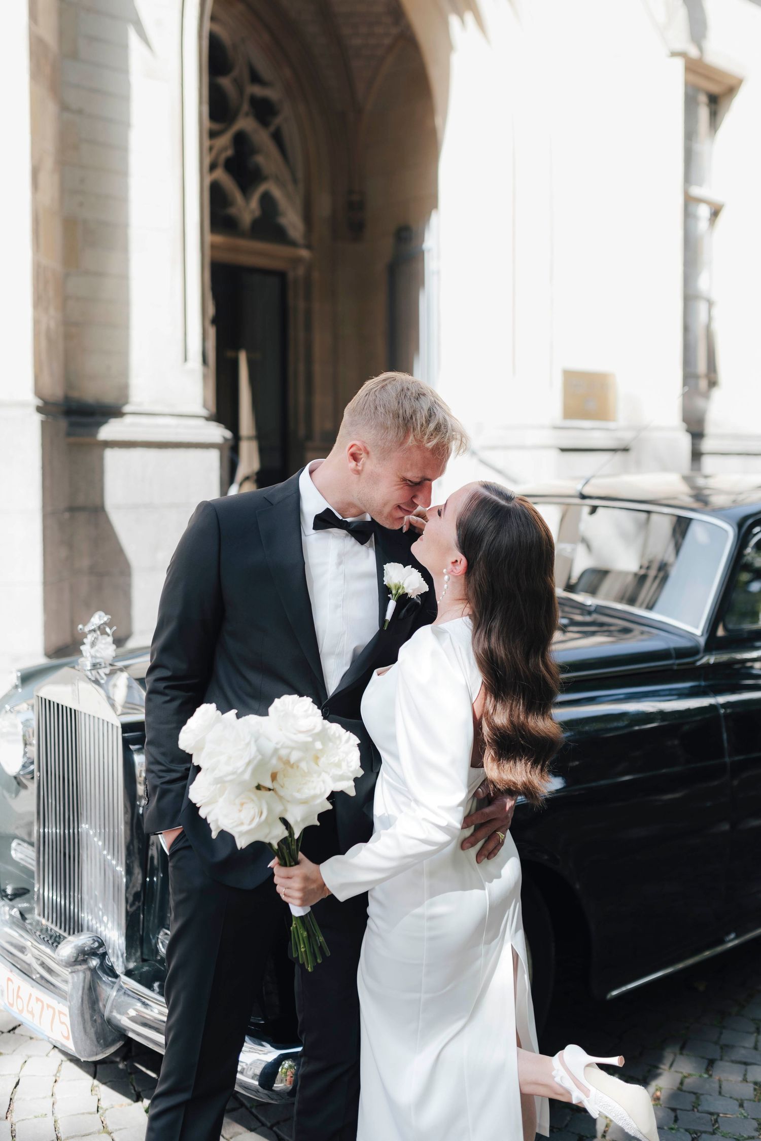 Bride and groom sharing a romantic moment beside a vintage black car outside a historic building on their wedding day.