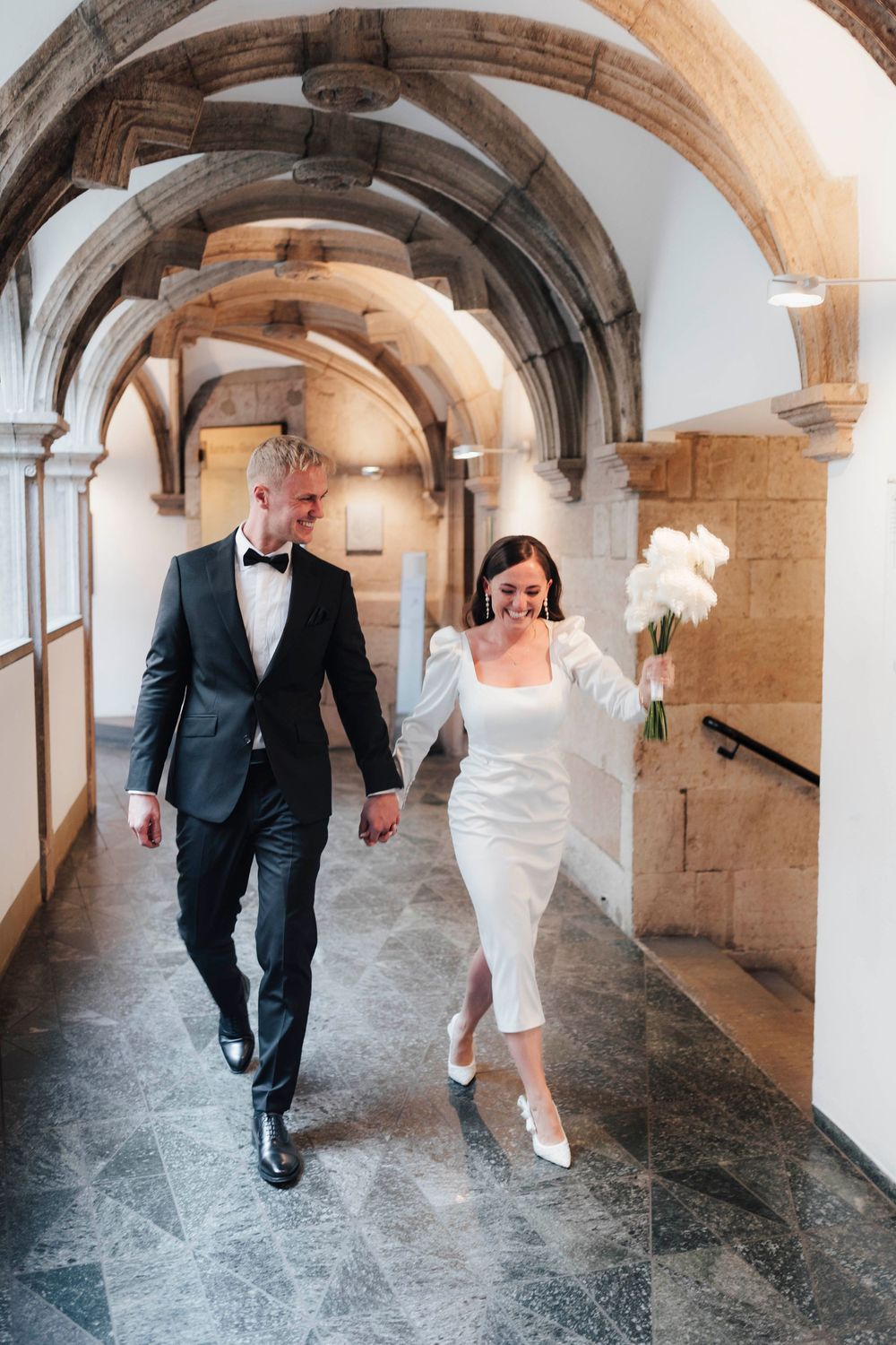 A bride and groom walk hand-in-hand through an arched stone corridor, she holds white tulips wearing a midi dress.