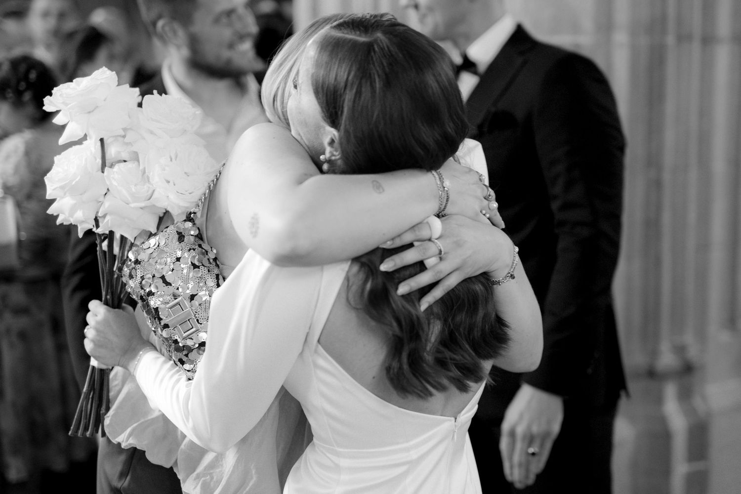 Black and white photo of a bride hugging a guest while holding a white rose bouquet at a wedding ceremony.
