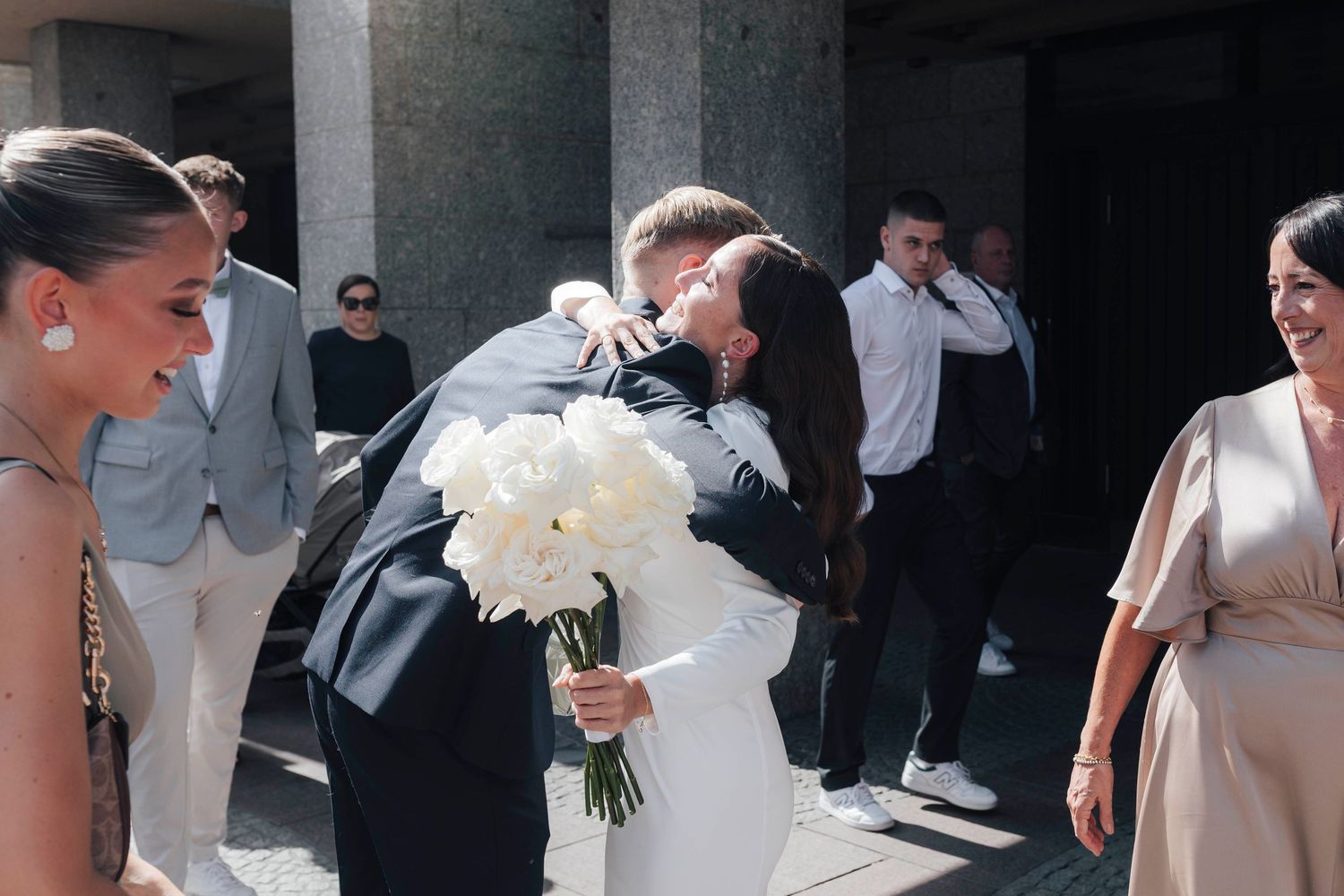 Bride and groom share a heartfelt embrace outside a wedding venue, holding white peony bouquet.
