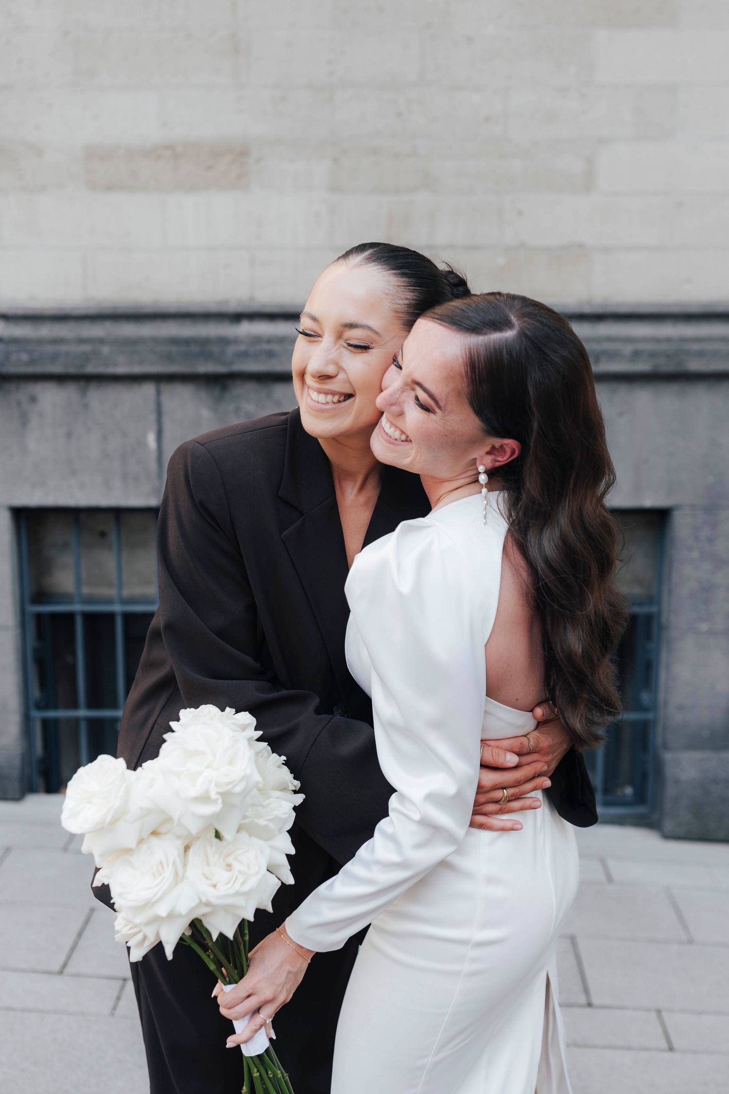 Two brides embracing outdoors, one in a white satin gown holding white peonies, the other in a black blazer, smiling joyfully.