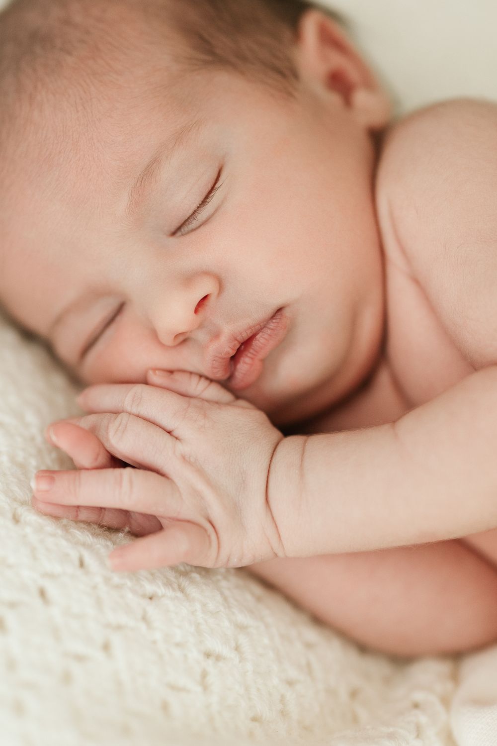 Peaceful newborn baby sleeping with hands clasped together on a soft cream knit blanket.