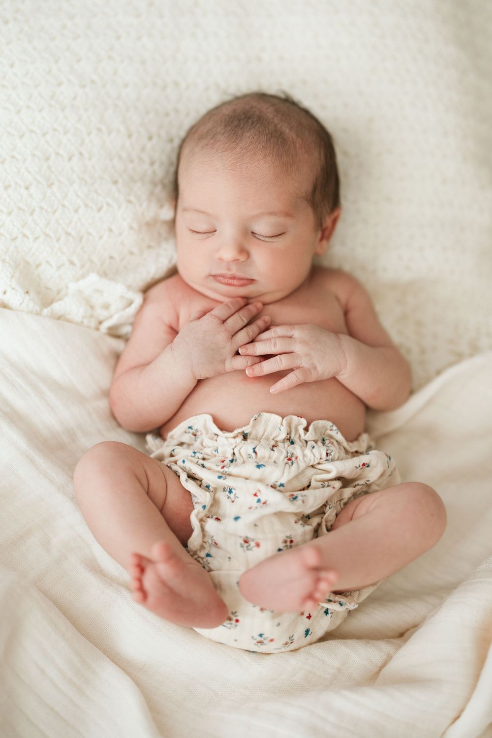 Peaceful newborn baby sleeping on cream knit blanket wearing floral bloomers with tiny hands folded on chest.