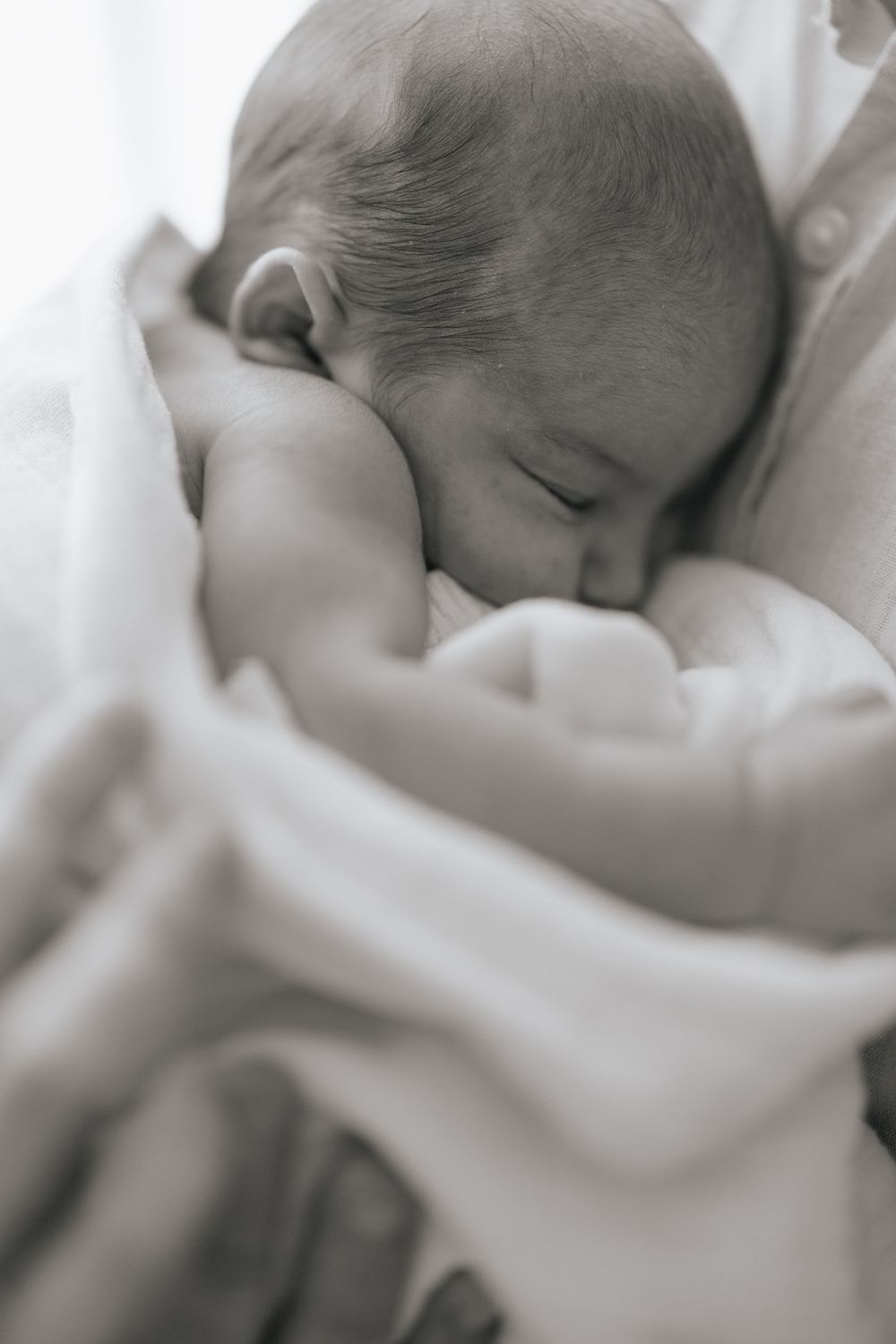 Newborn baby sleeping peacefully wrapped in a soft white blanket cradled in loving arms, black and white photo.