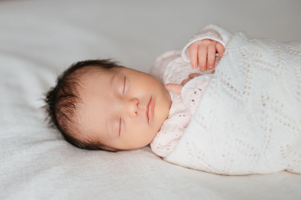 Peaceful newborn baby sleeping swaddled in a soft white knit blanket with dark hair and tiny hands visible.