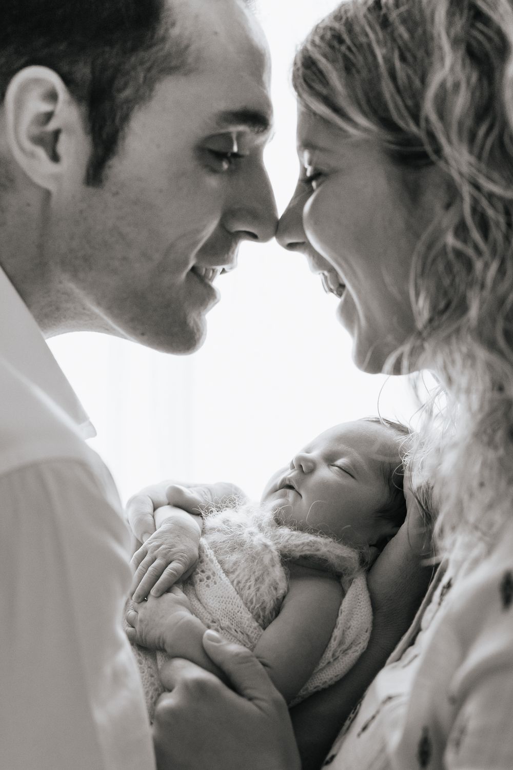 Black and white photo of smiling parents touching noses while cradling their sleeping newborn baby.