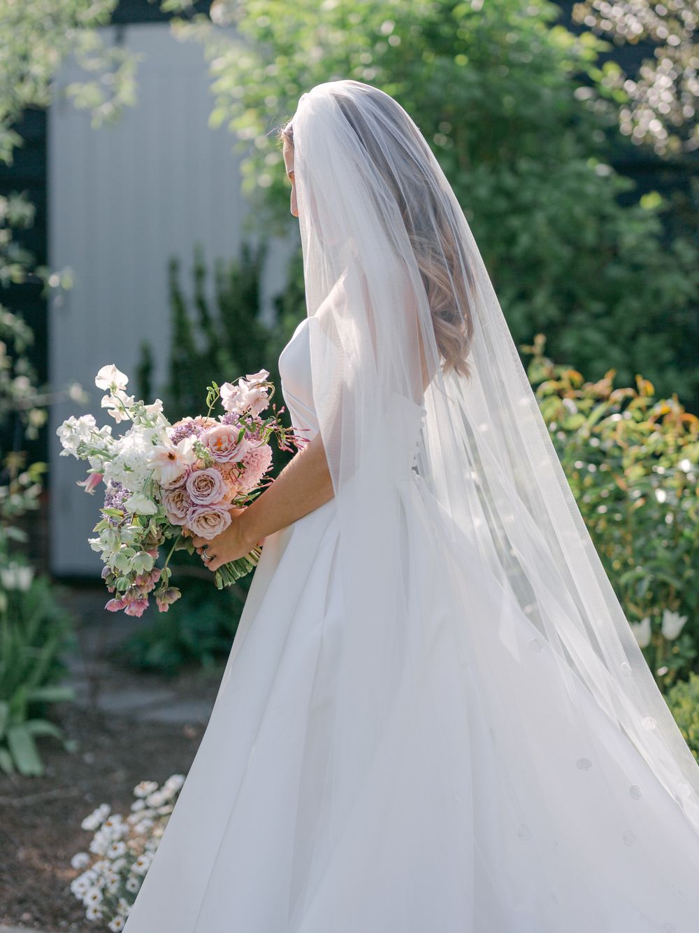 Bride in flowing veil holding delicate pink and white bouquet in garden setting.