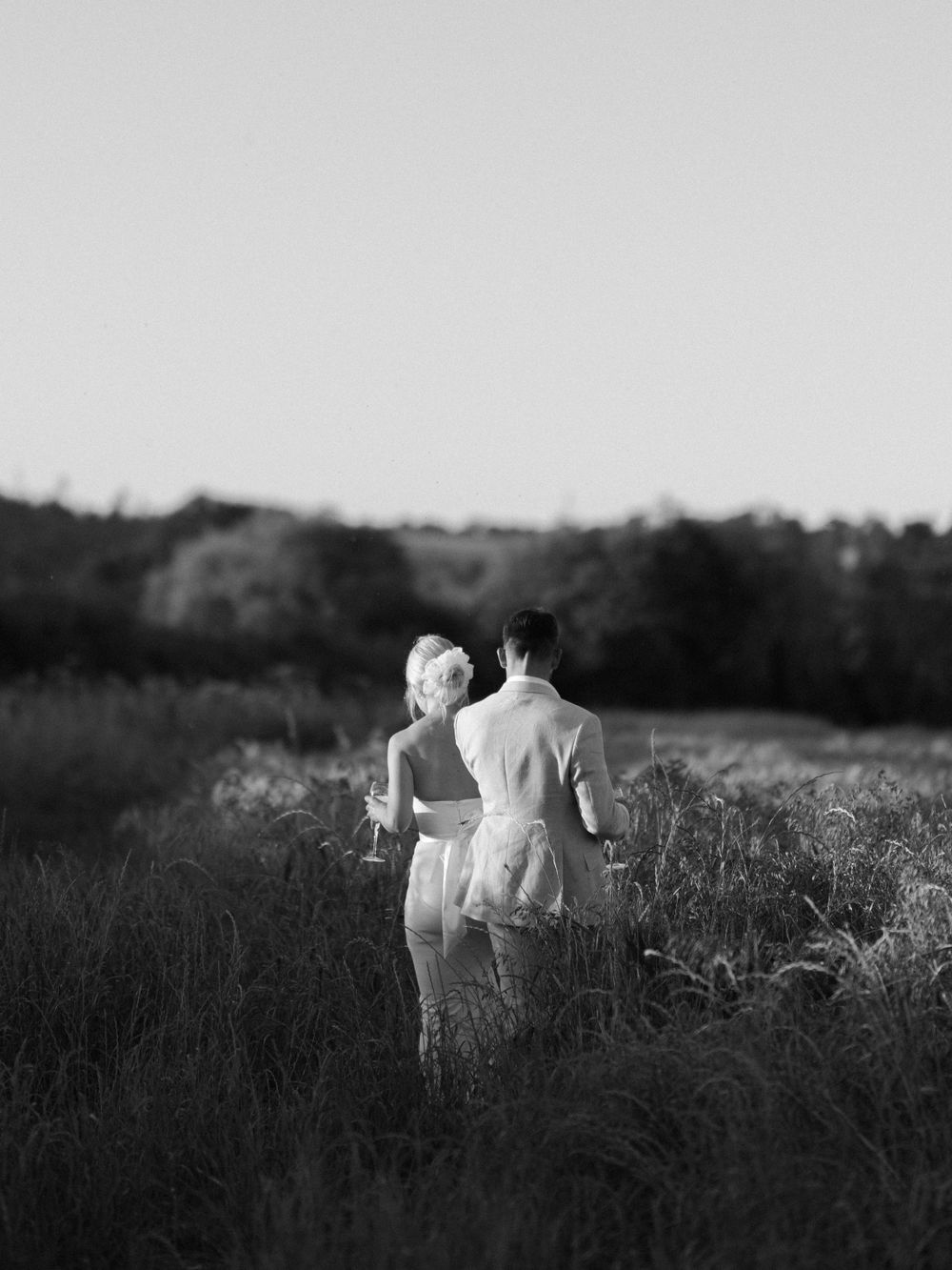 Black and white photo of a couple walking through tall grass at sunset.