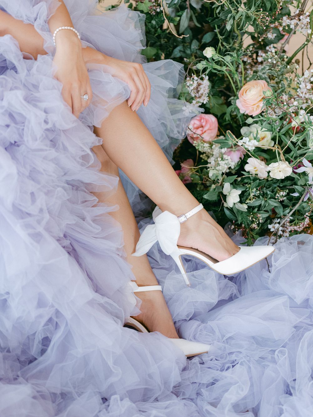 White bridal heels and flowing lavender tulle dress next to pink floral arrangements.
