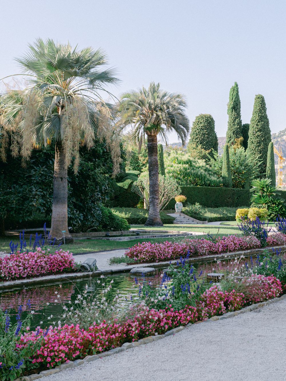 Formal garden path lined with palm trees and vibrant pink flowers alongside reflecting pools.