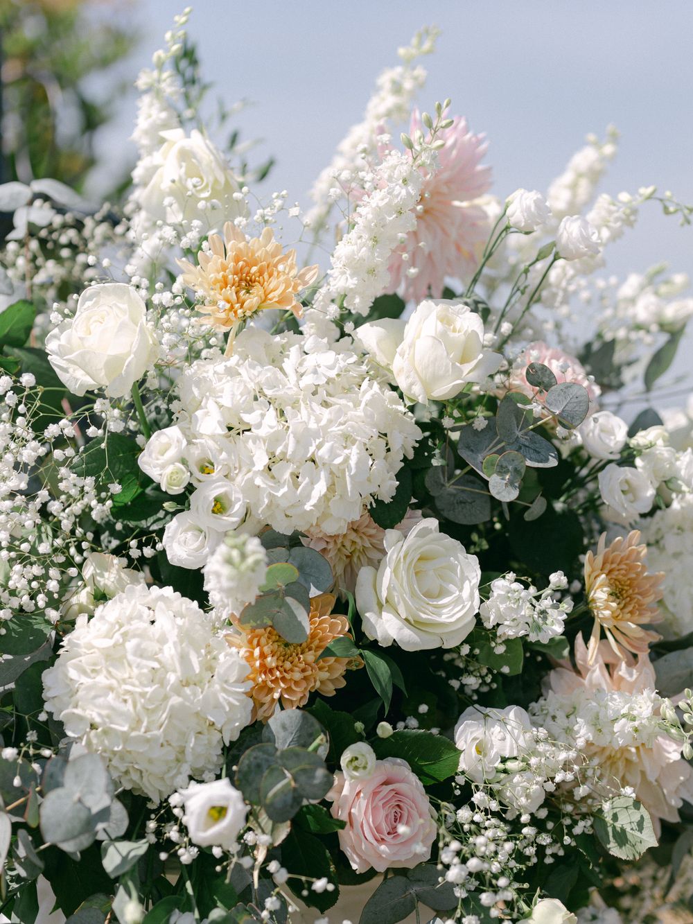 Elegant white floral arrangement with roses, peonies, and baby's breath against a soft blue background.