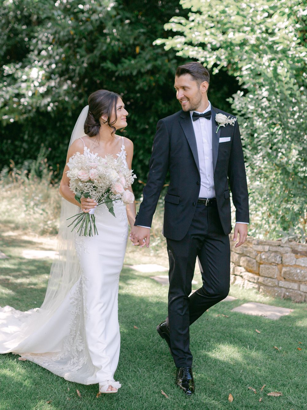 A couple in wedding attire walking and holding hands in a sunny garden setting.