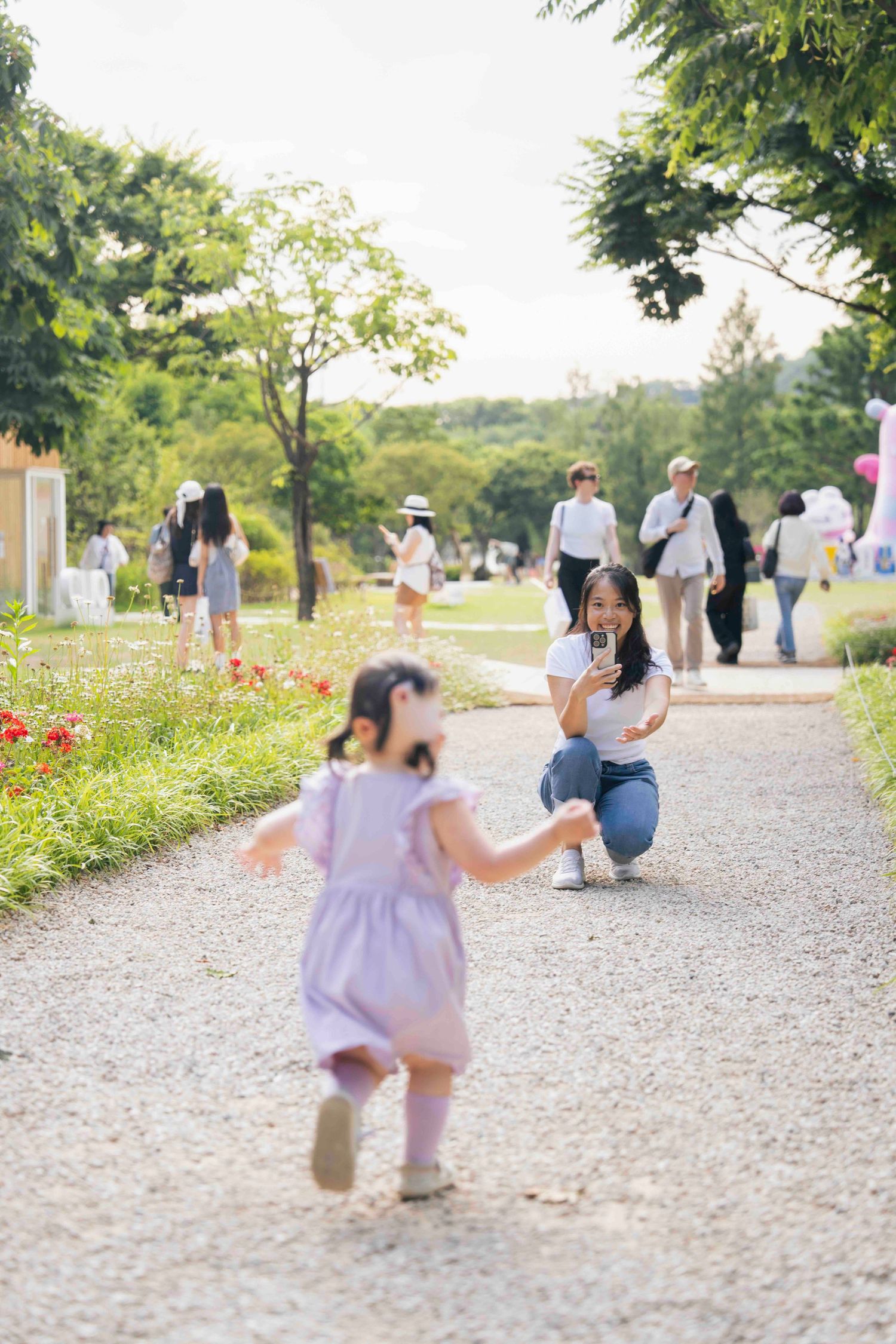 A mother crouches to photograph her young daughter running toward her on a park path surrounded by greenery.