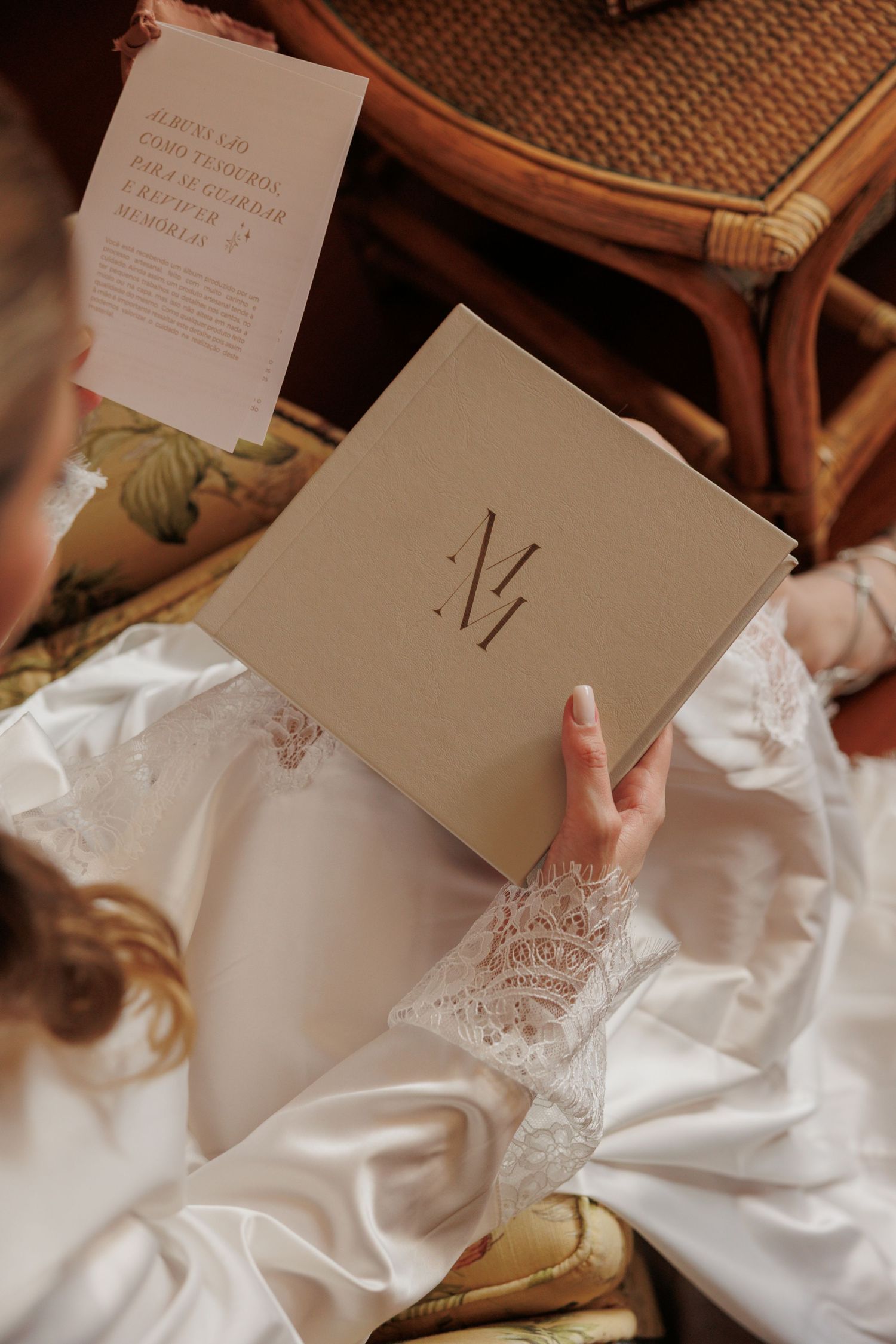 Bride in white lace robe reading a kraft paper booklet with monogram M initial, wedding morning preparation.