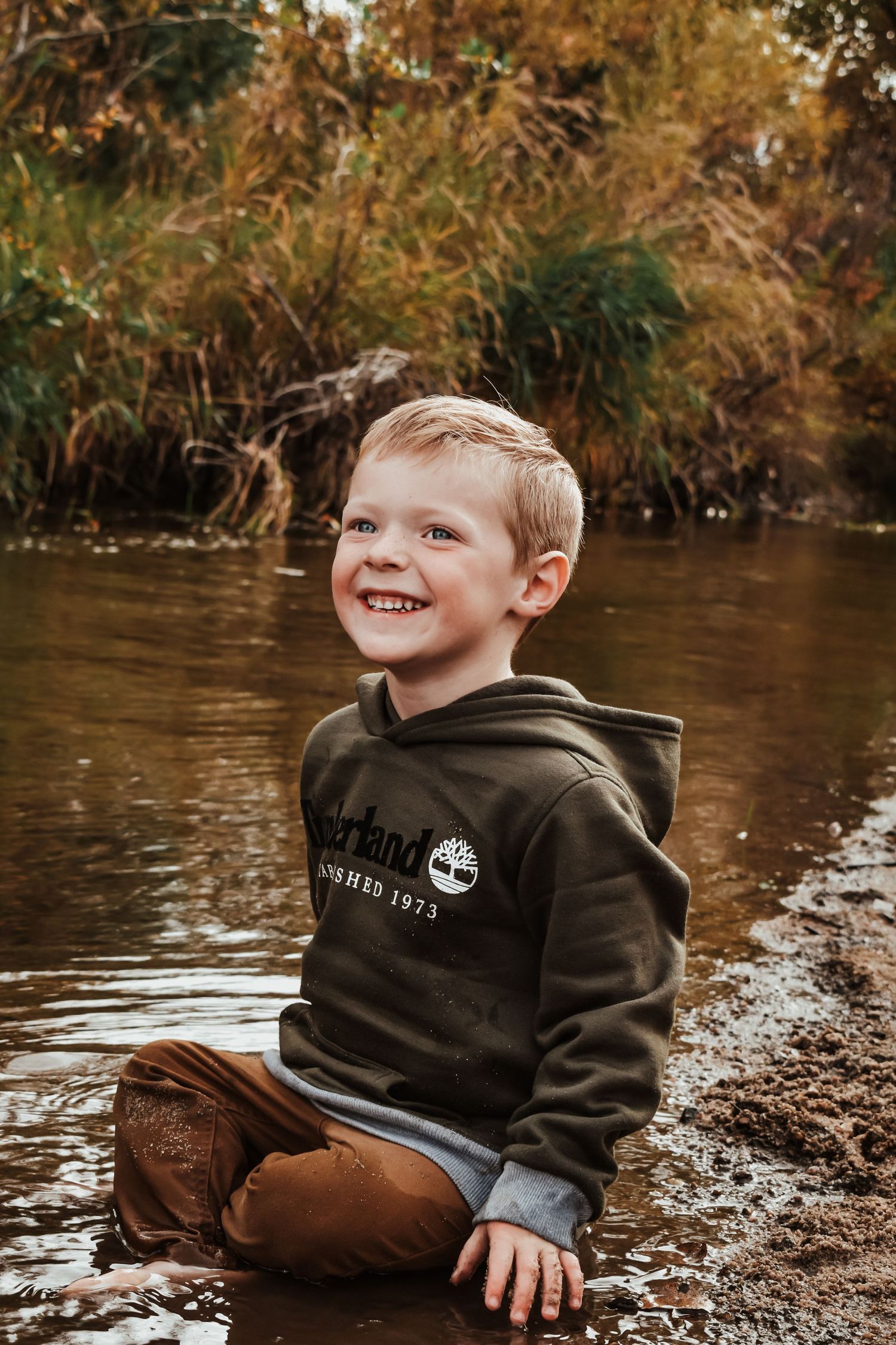Smiling young boy sitting in shallow water wearing a Timberland hoodie, surrounded by autumn foliage.