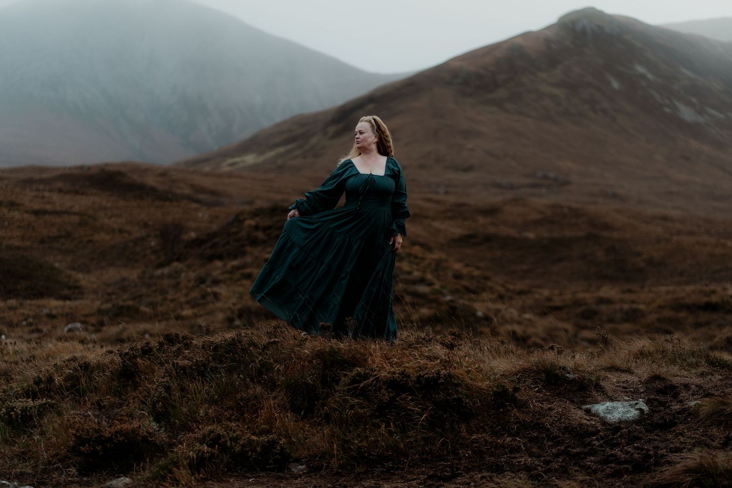 Woman in green medieval gown standing on misty Scottish highlands with dramatic mountain backdrop.