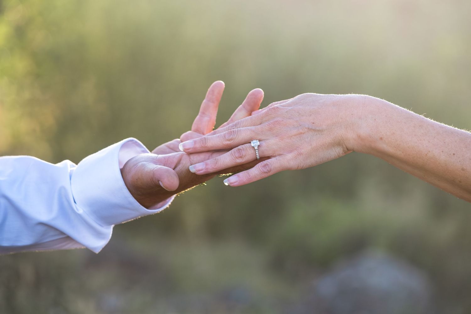 Close-up of a couple tenderly touching hands. On her hand is a diamond engagement ring in the outdoors with a soft green bokeh background.
