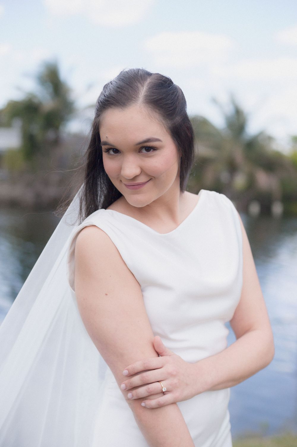 Bride in white sleeveless gown and veil smiles gracefully outdoors near a serene waterway with palm trees.