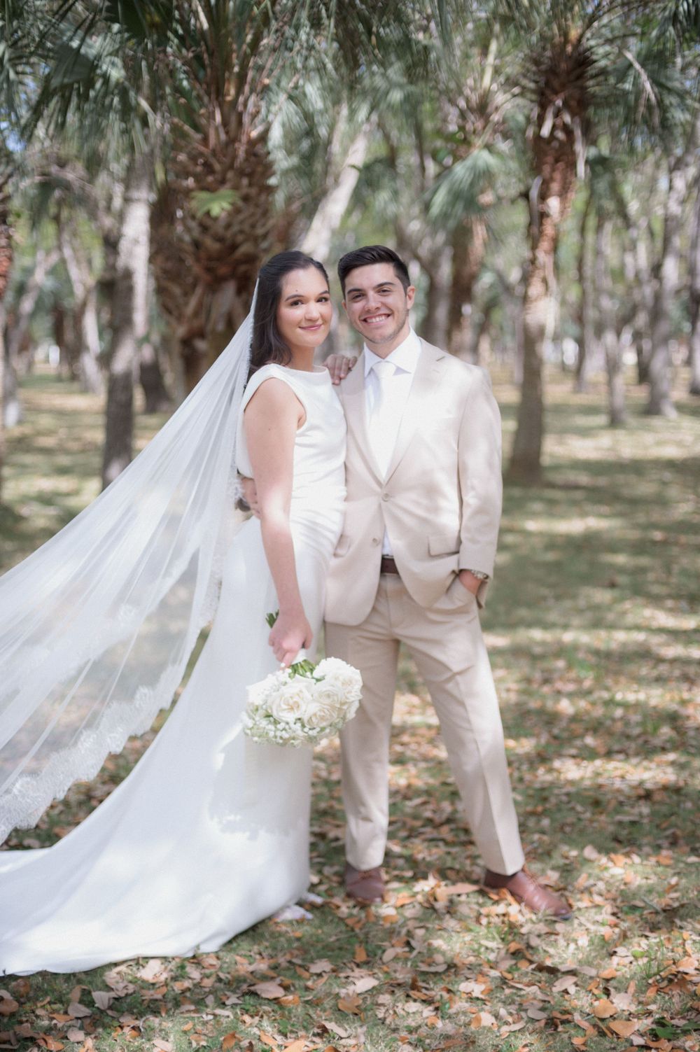 Bride and groom pose together outdoors among palm trees, she in a white gown with veil, he in a beige suit.