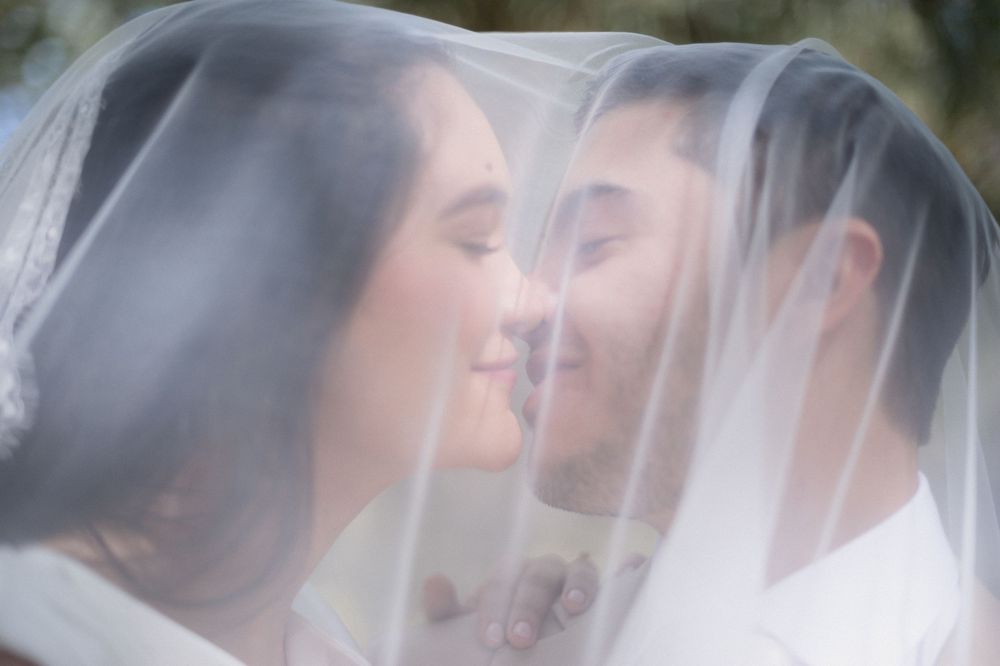 Bride and groom sharing a romantic moment under a wedding veil, faces close together in a tender almost-kiss.