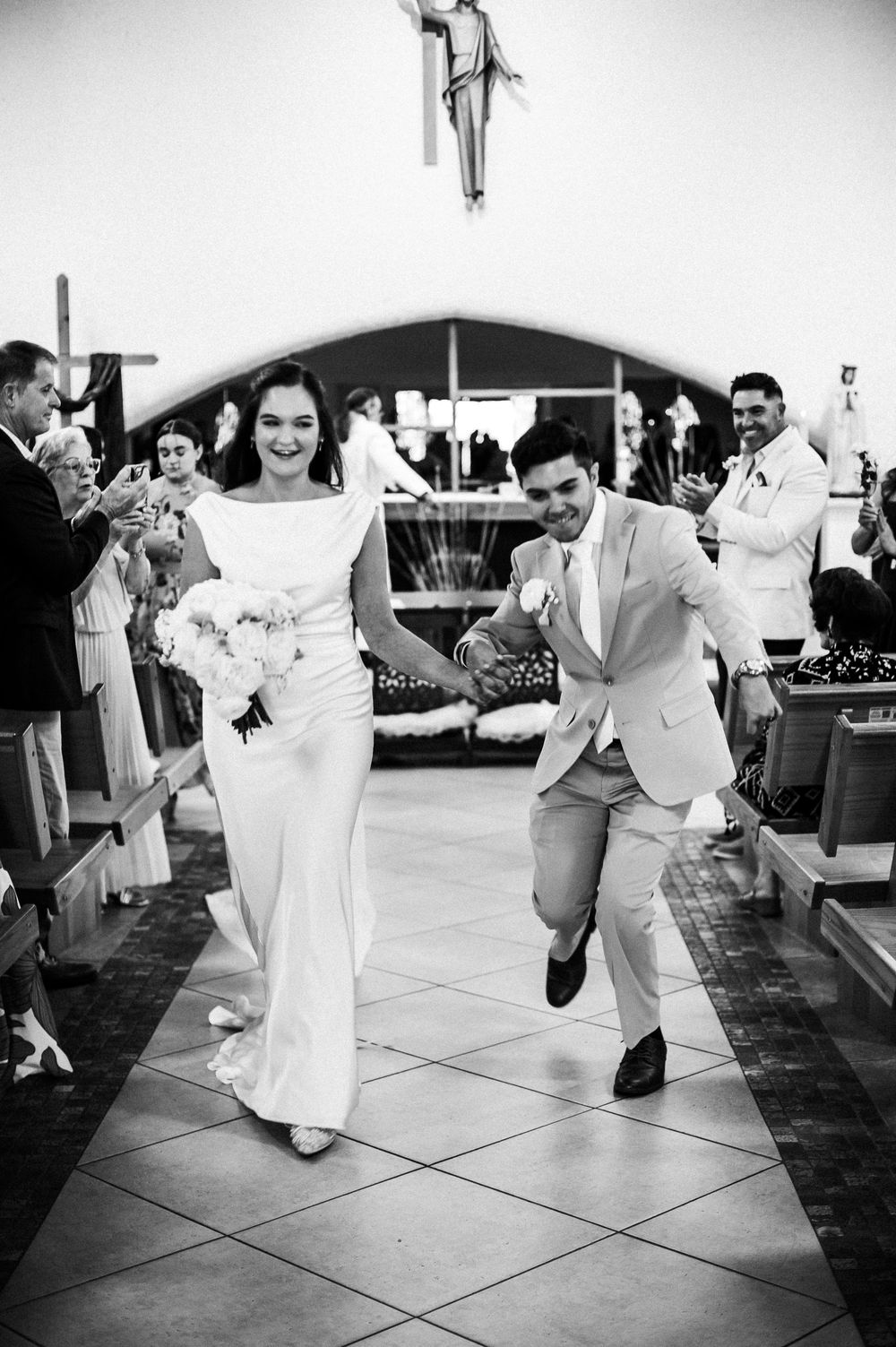 Joyful newlyweds run down church aisle after wedding ceremony, bride in white gown holding bouquet, groom in light suit.