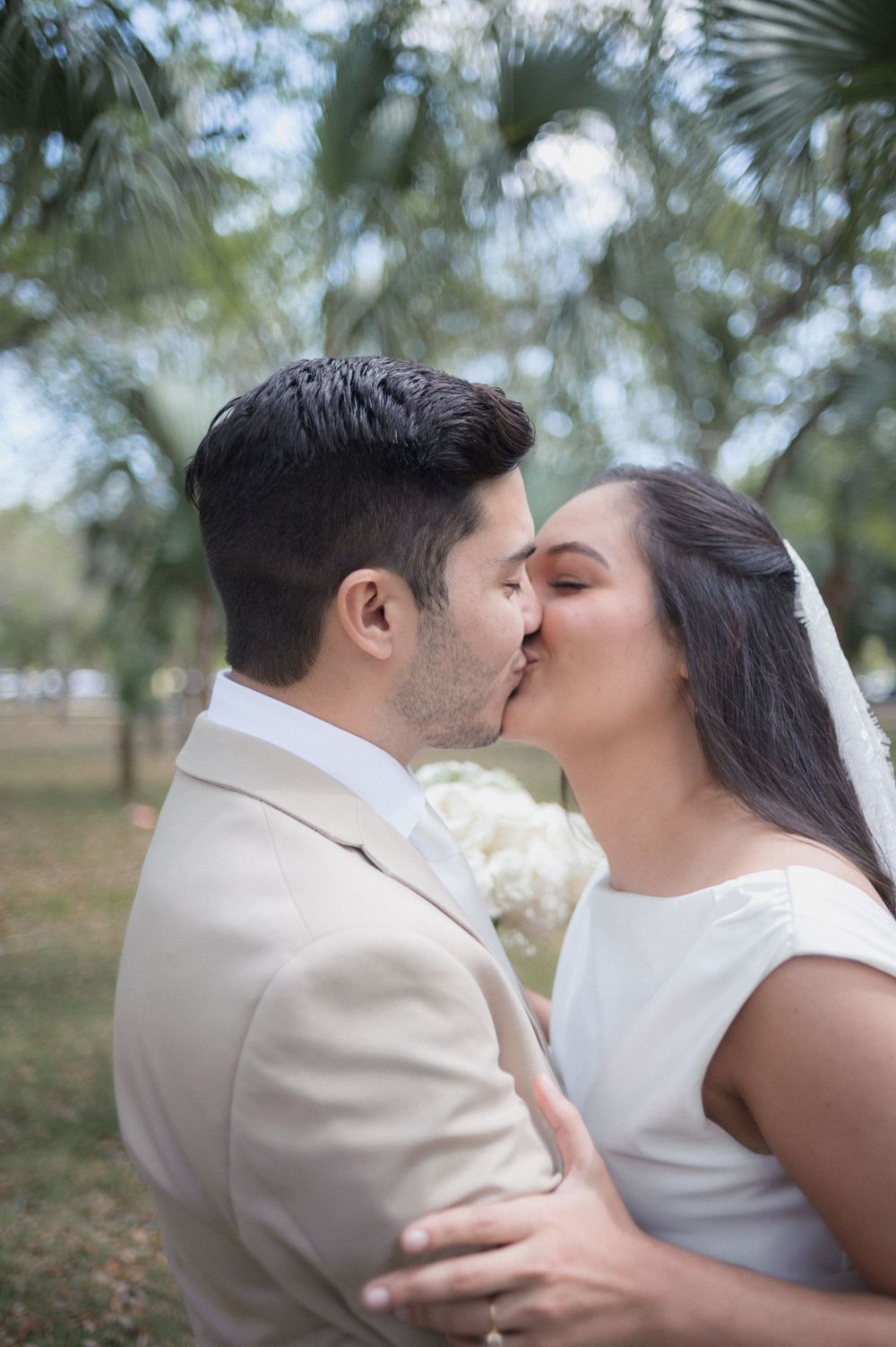 A bride and groom share a romantic kiss outdoors, surrounded by lush green trees, as the bride holds a white floral bouquet.