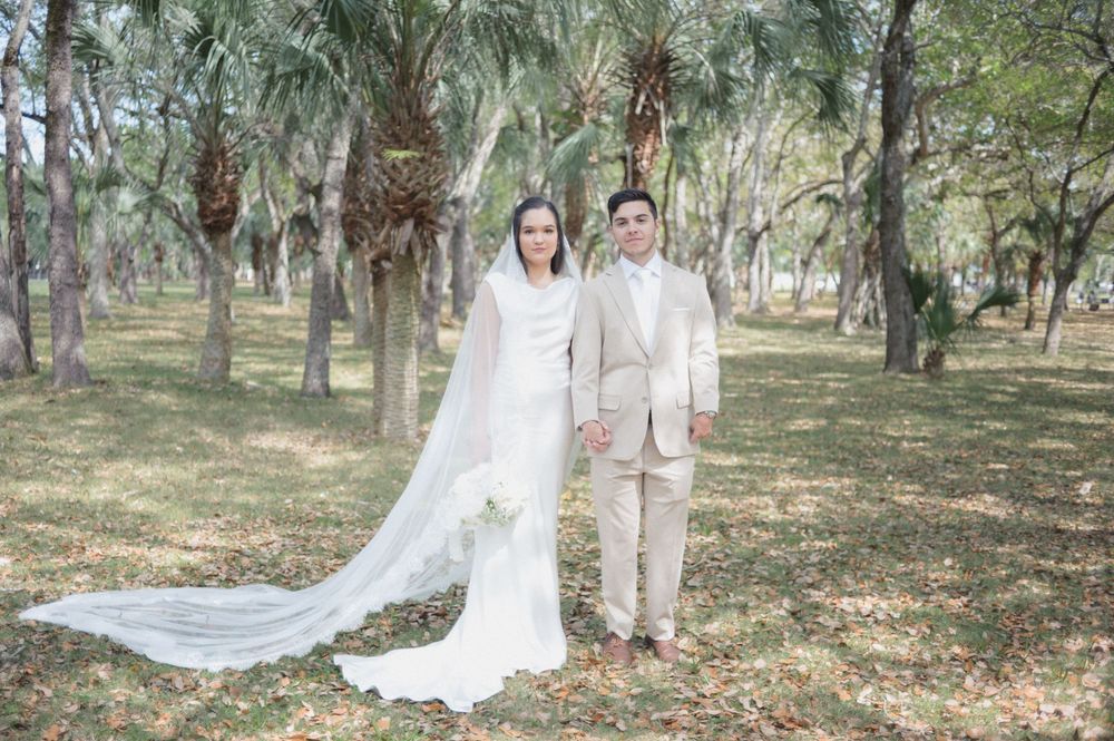 Bride in white gown with long veil and groom in beige suit holding hands in a lush green forest setting.