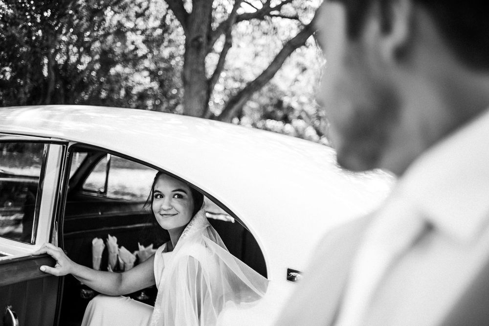 Black and white photo of a smiling bride in a wedding dress stepping out of a vintage car, greeted by a groom.