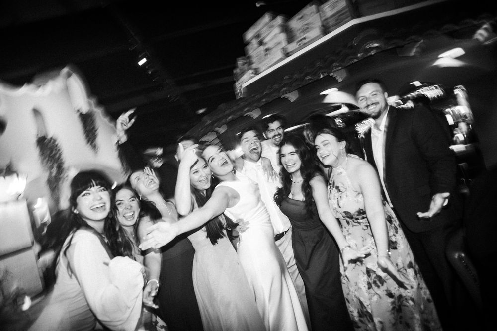 Black and white photo of a joyful wedding party group selfie at a reception venue.