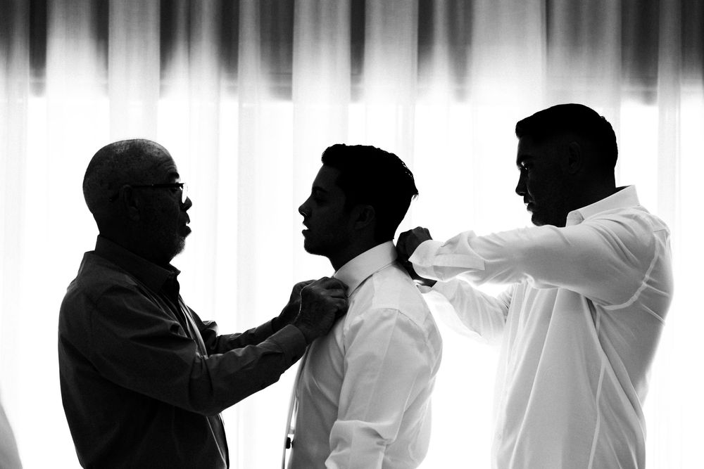 Black and white photo of a groom being helped with his tie by two men before a wedding ceremony.