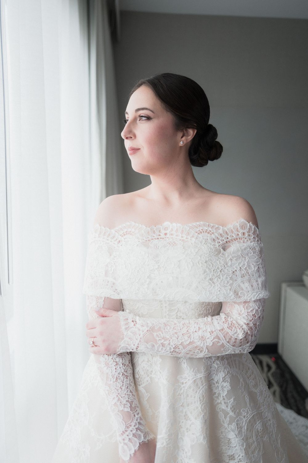 Elegant bride in off-shoulder lace wedding dress with long sleeves, hair in updo, gazing thoughtfully out a window.