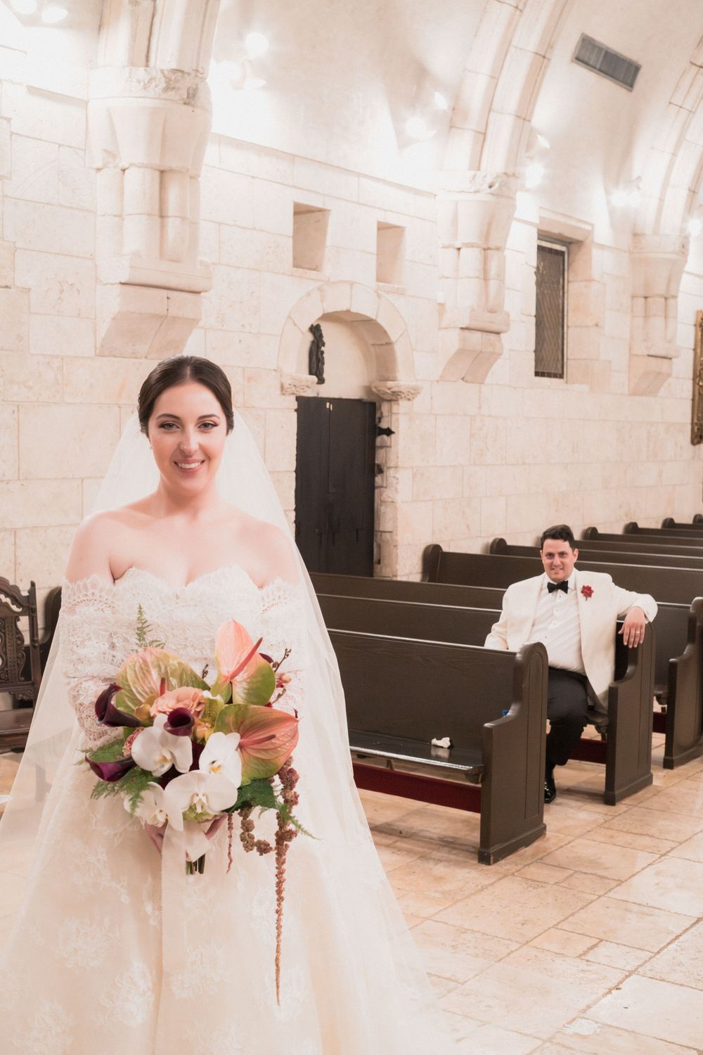 Bride in white gown holding floral bouquet stands in stone church aisle, groom in white suit seated on pew behind her.