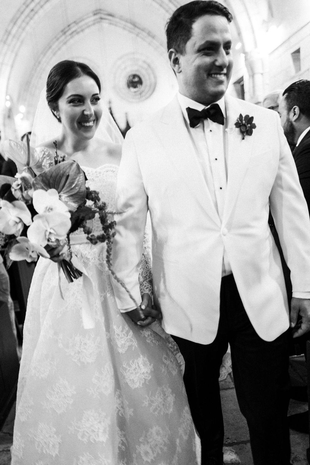 Black and white photo of a smiling bride and groom holding hands walking down the aisle after their wedding ceremony.