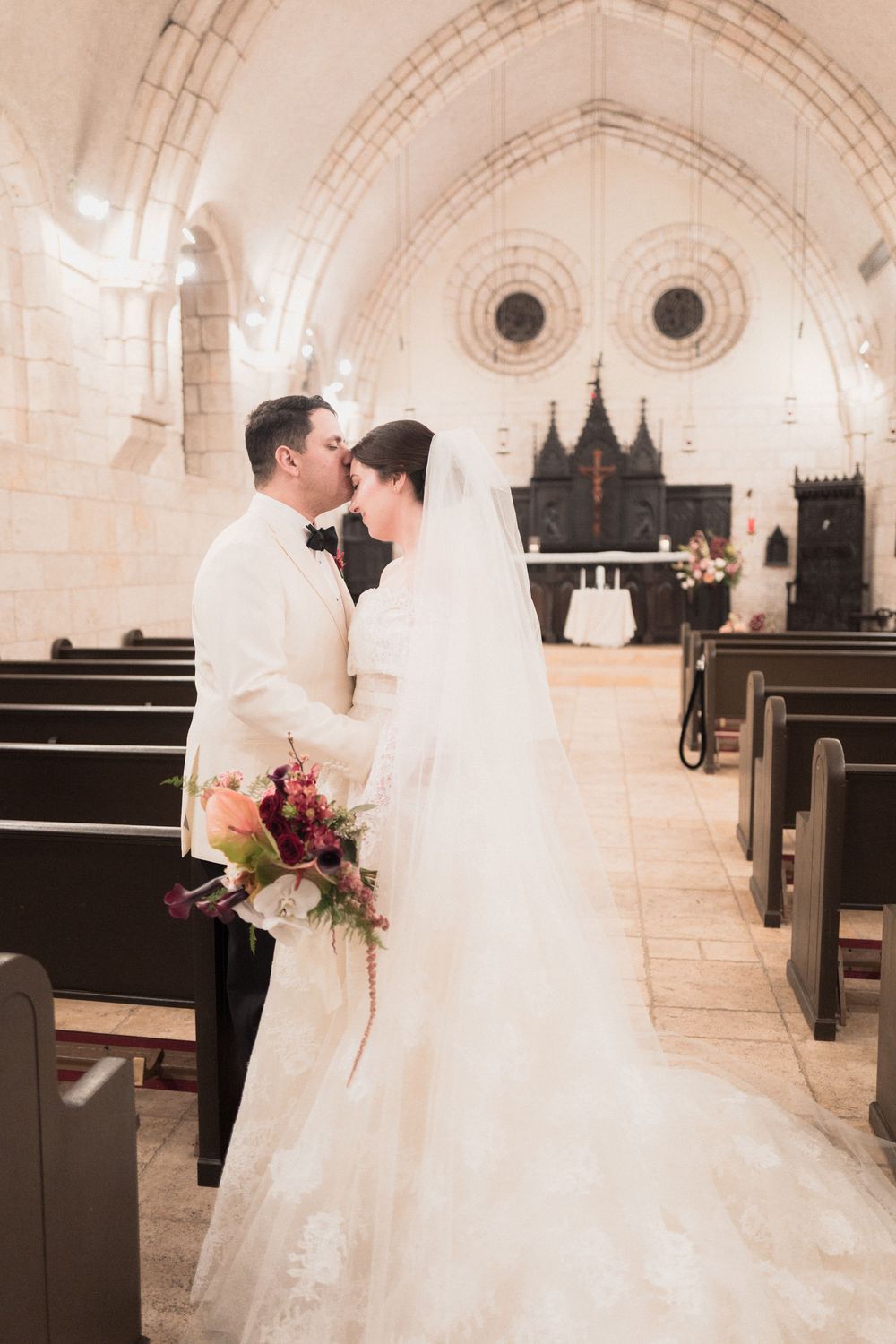 Bride and groom share a kiss in a stunning stone church, bride holding a vibrant bouquet in a lace wedding gown.