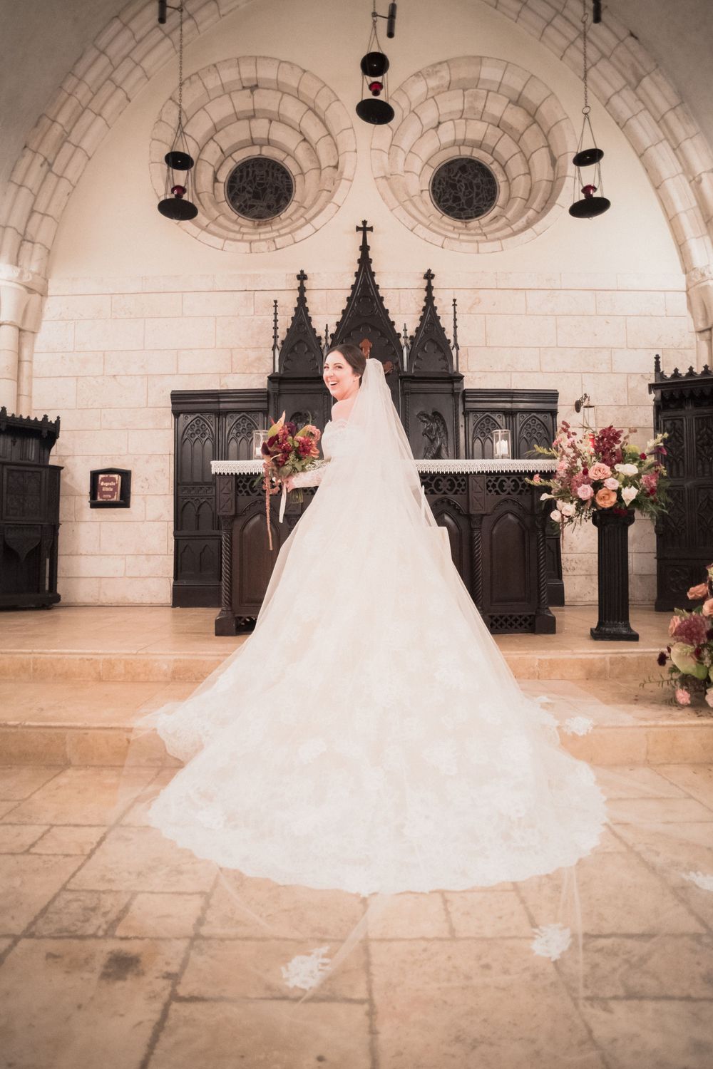 Bride in lace gown with cathedral veil standing before gothic altar in stone chapel with floral decor.