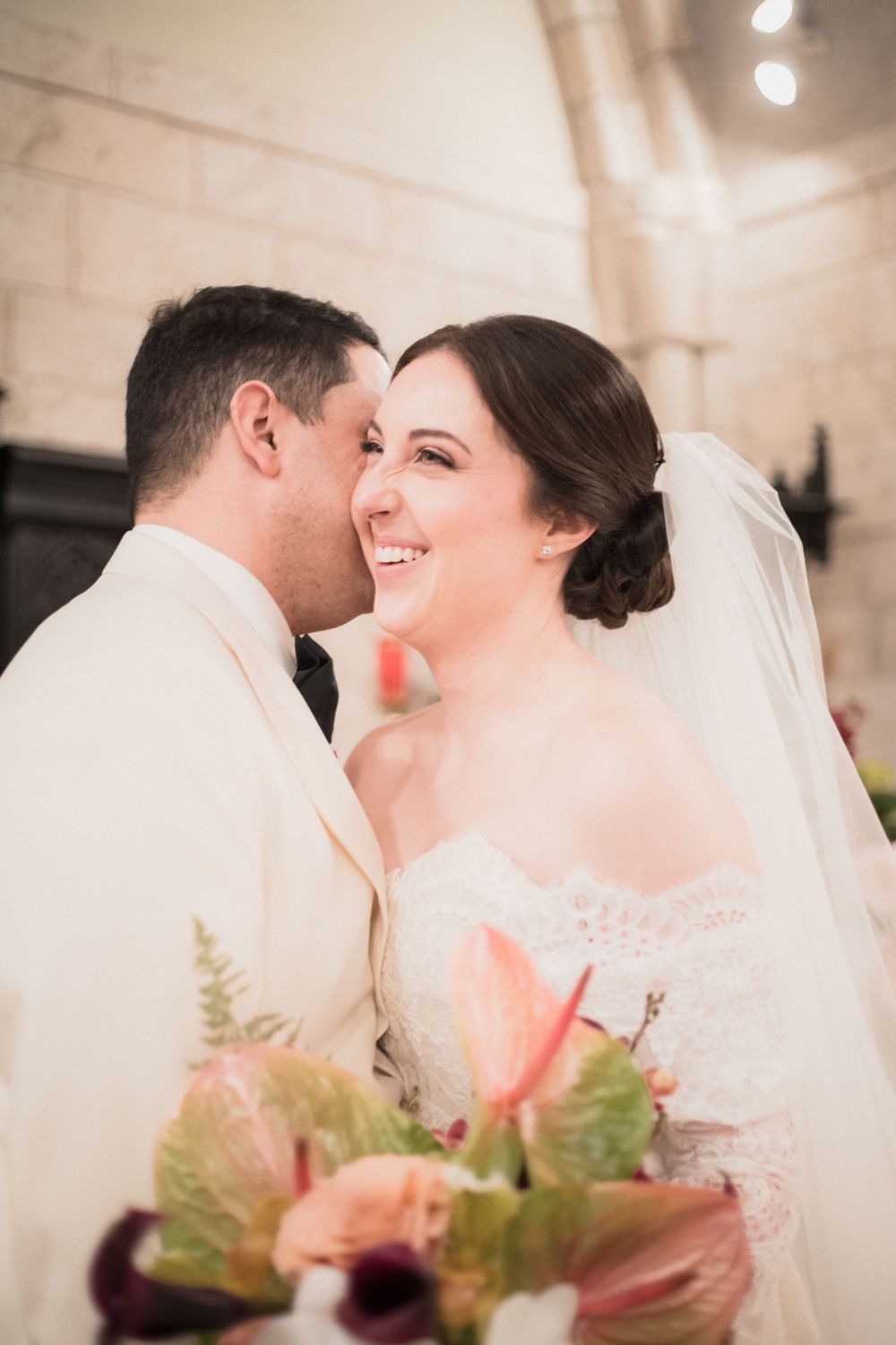Smiling bride in white lace gown and veil holds pink bouquet as groom in ivory tuxedo whispers in her ear.