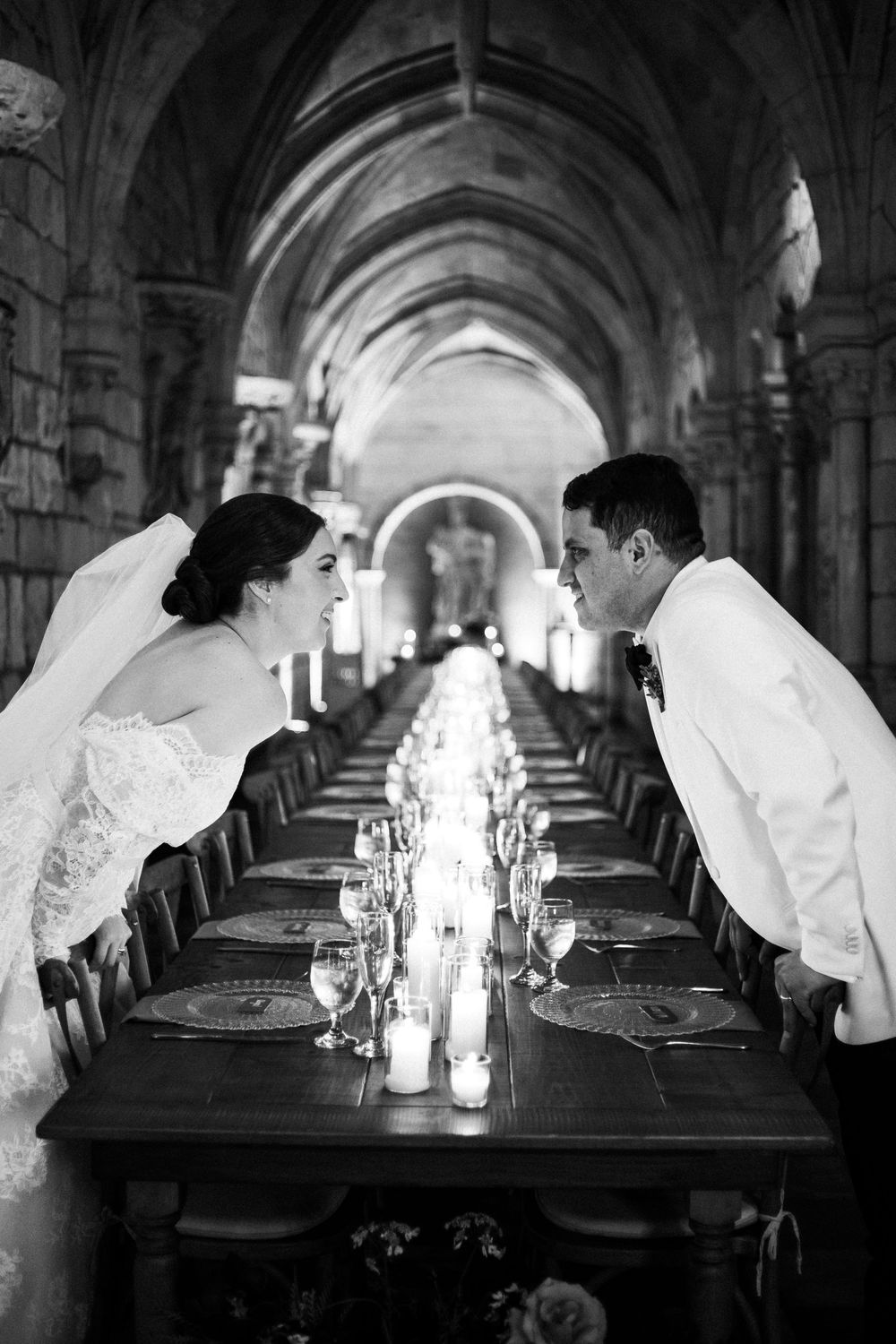 Black and white photo of bride and groom leaning toward each other across an elegant candlelit table in a Gothic archway.