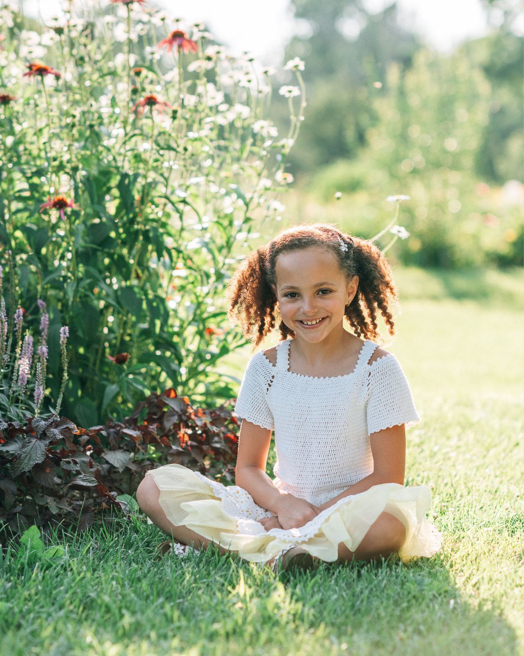 Young girl with curly hair sits cross-legged on grass beside colorful wildflowers, wearing white top and yellow skirt.
