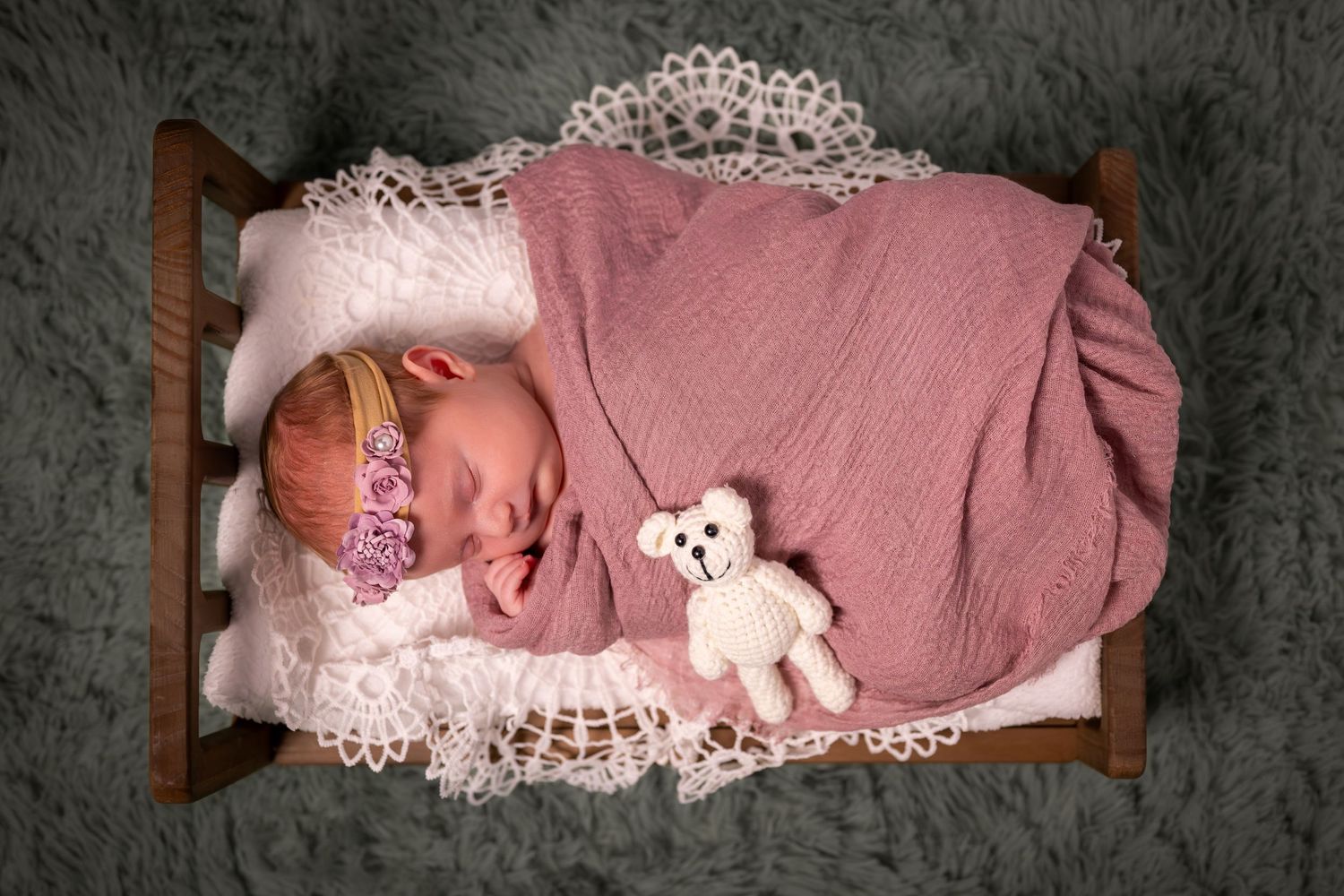 Newborn baby sleeping in tiny wooden bed with pink wrap, lace blanket, floral headband, and small knit teddy bear.
