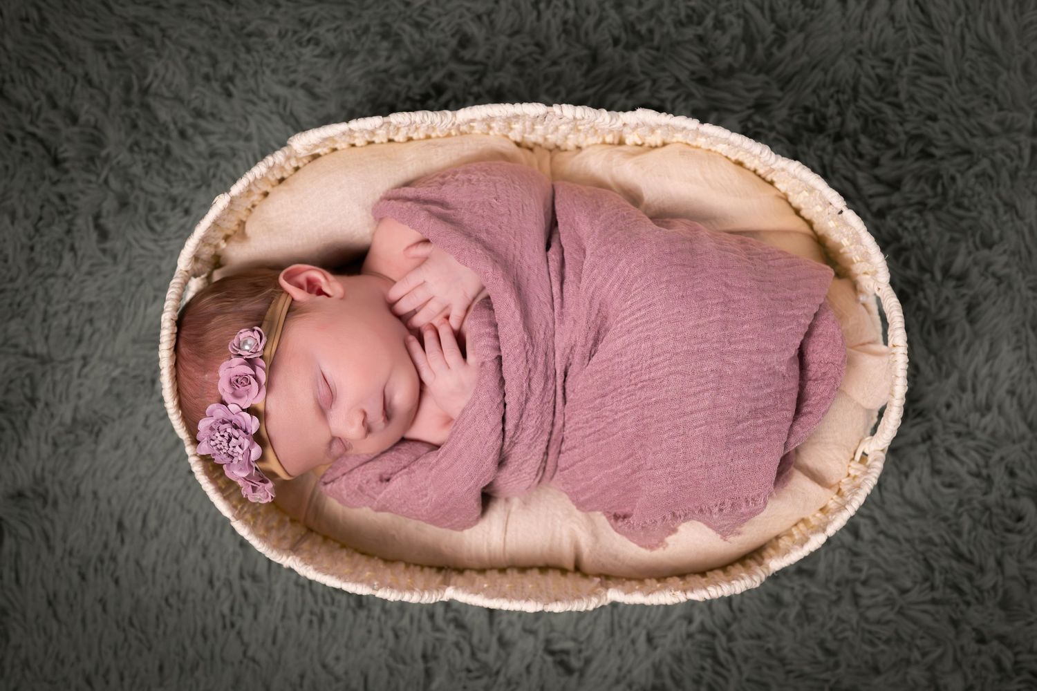 Newborn baby swaddled in pink wrap with floral headband sleeping peacefully in a wicker basket on gray fur.