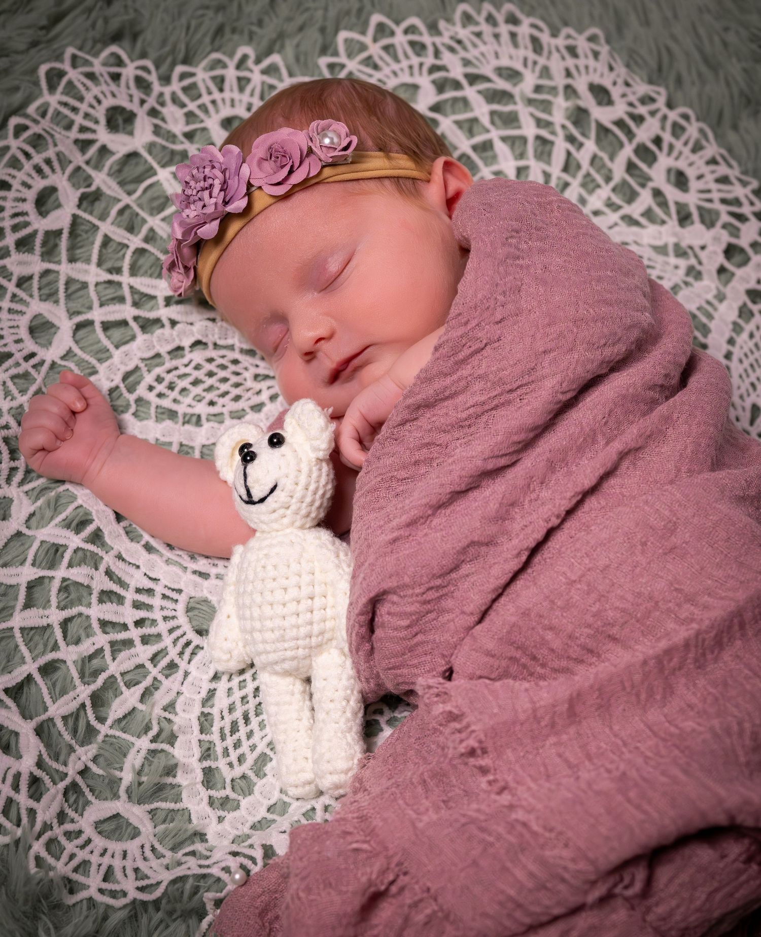 Sleeping newborn baby wrapped in pink blanket with floral headband cuddling a white crocheted teddy bear.