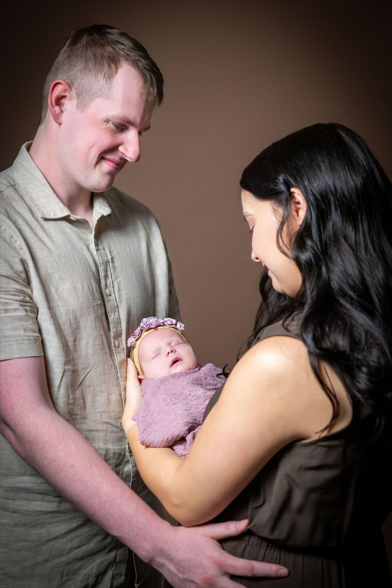 A couple lovingly gazes at their sleeping newborn baby wrapped in purple, in a studio portrait.