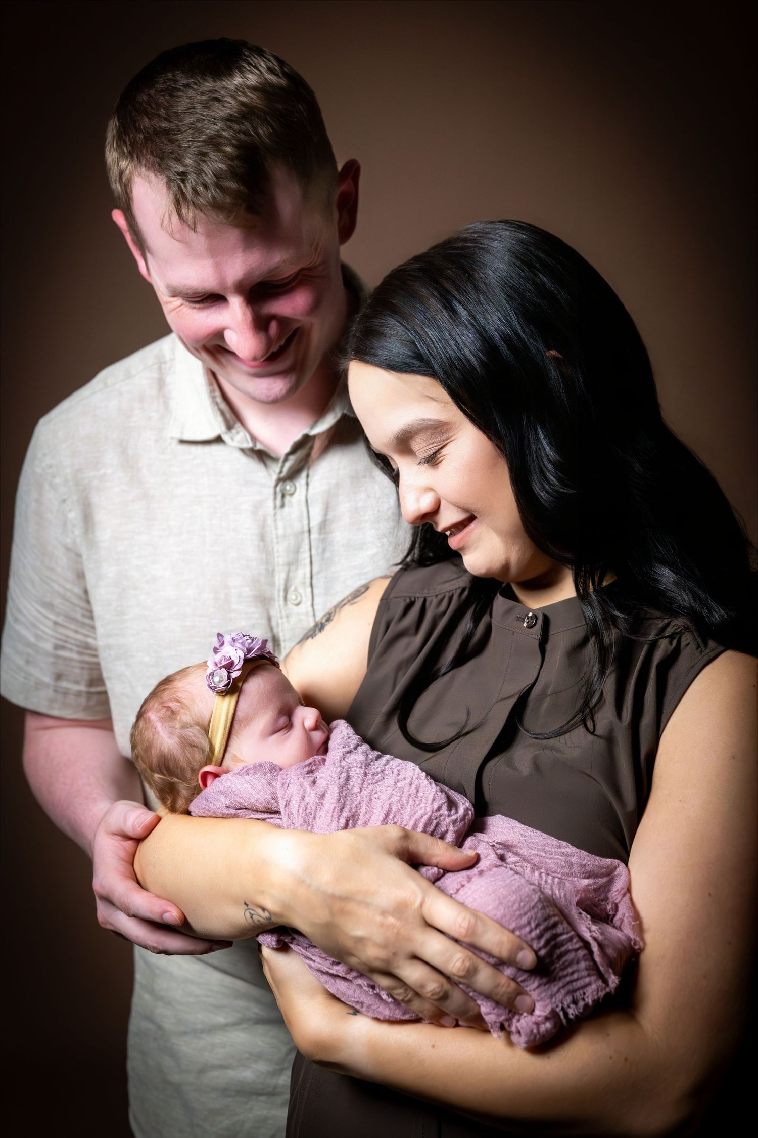 Smiling parents lovingly hold their newborn baby wrapped in purple, posing for a portrait photo.
