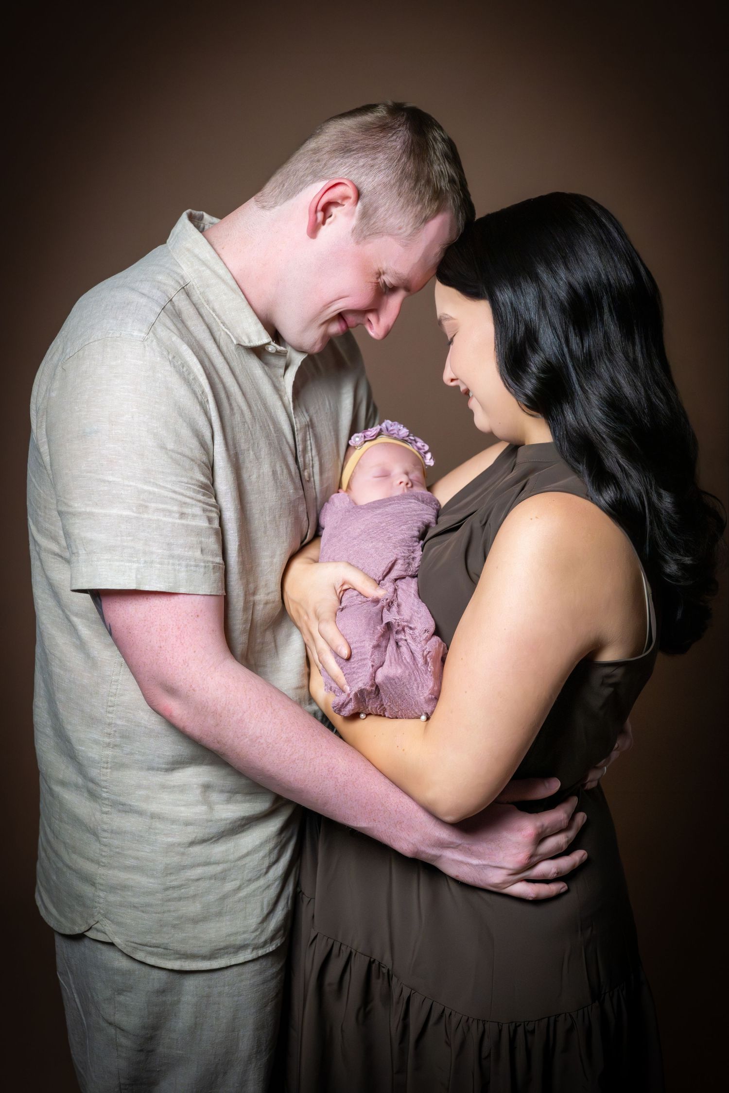 Couple touching foreheads tenderly while holding their purple-swaddled newborn in a studio portrait.