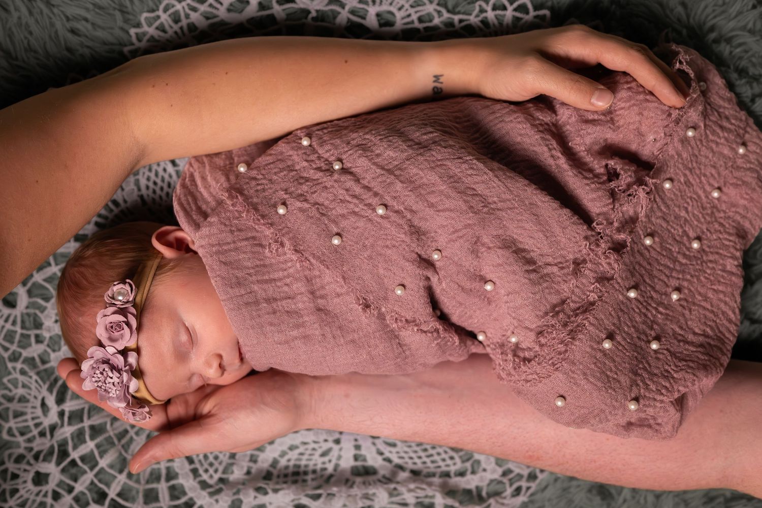 Newborn baby sleeping peacefully swaddled in a pink muslin wrap with pearl details and floral headband.